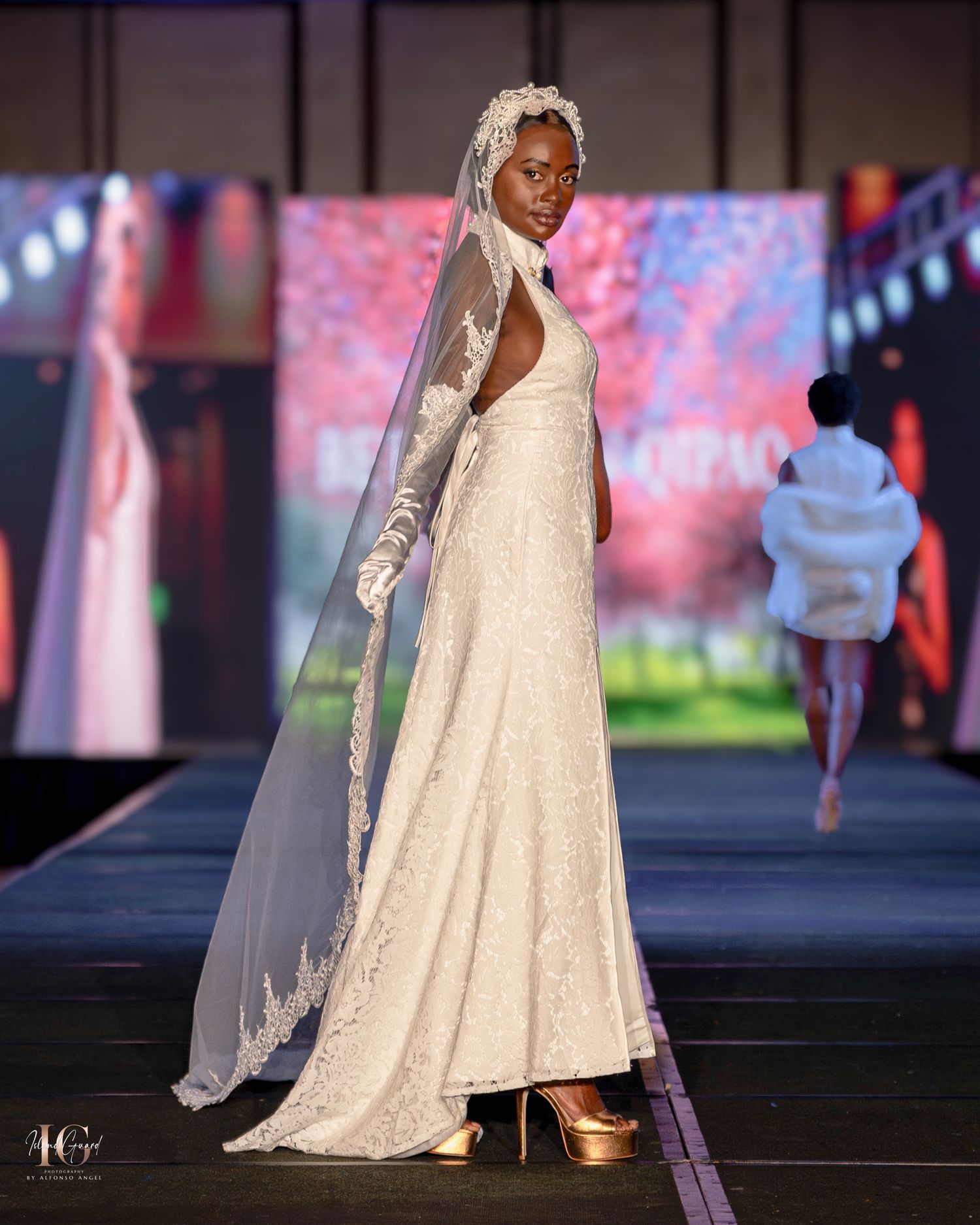 Elegant white lace wedding gown displayed during evening fashion show with colorful backdrop.