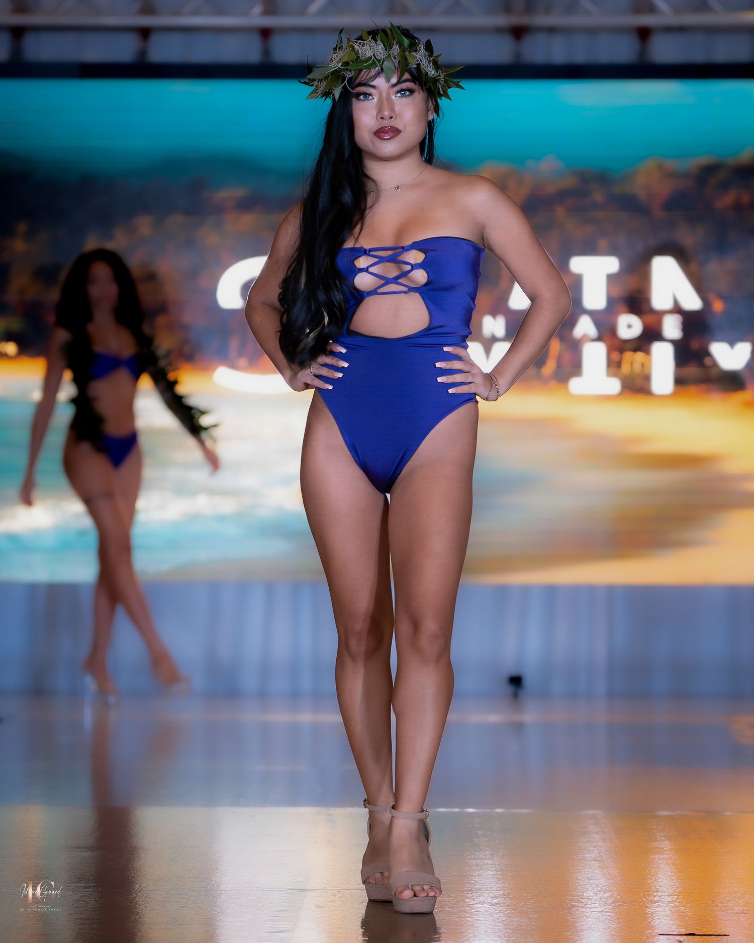 Model presents navy blue one-piece swimsuit on catwalk with ocean-themed lighting.