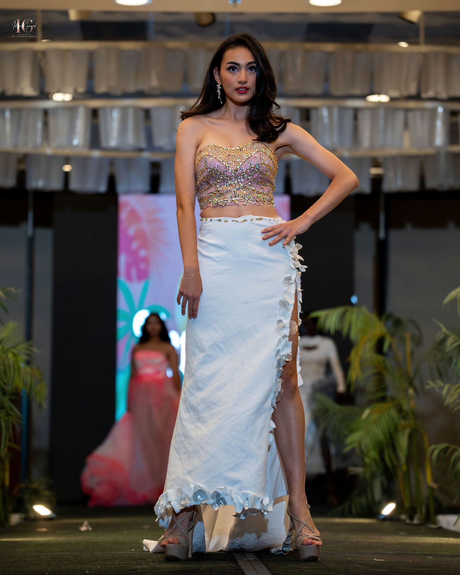 Model walks runway in sparkly strapless crop top and white ruffled maxi skirt with side slit at fashion show.