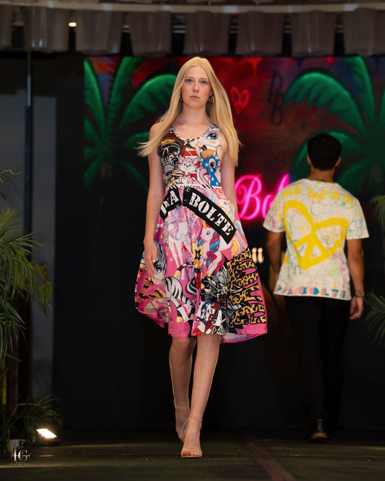 Model walks runway in vibrant floral print romper during fashion show with colorful neon lighting and dark backdrop.