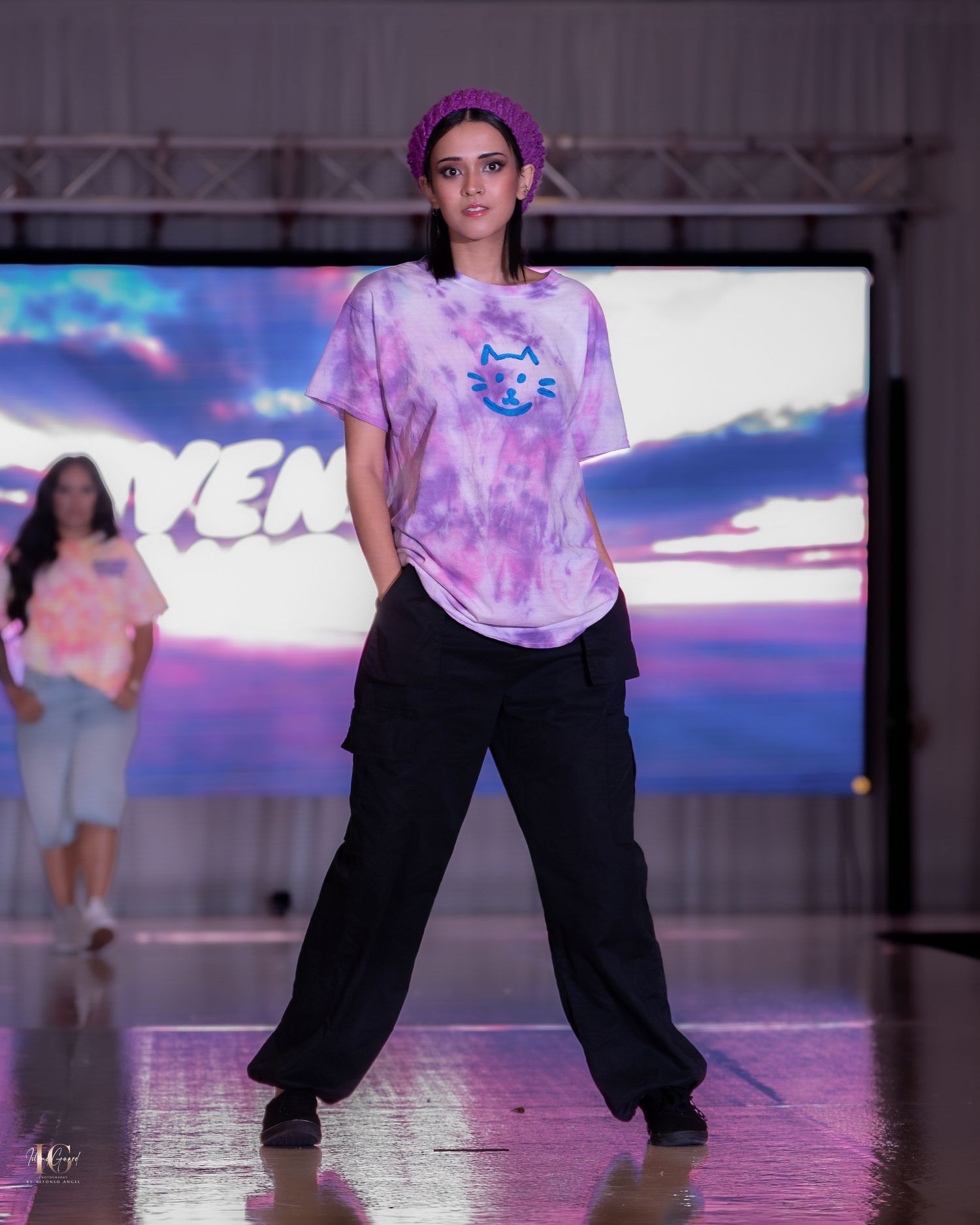 A dancer in a tie-dye shirt and black pants performs on a reflective stage with colorful lighting effects.