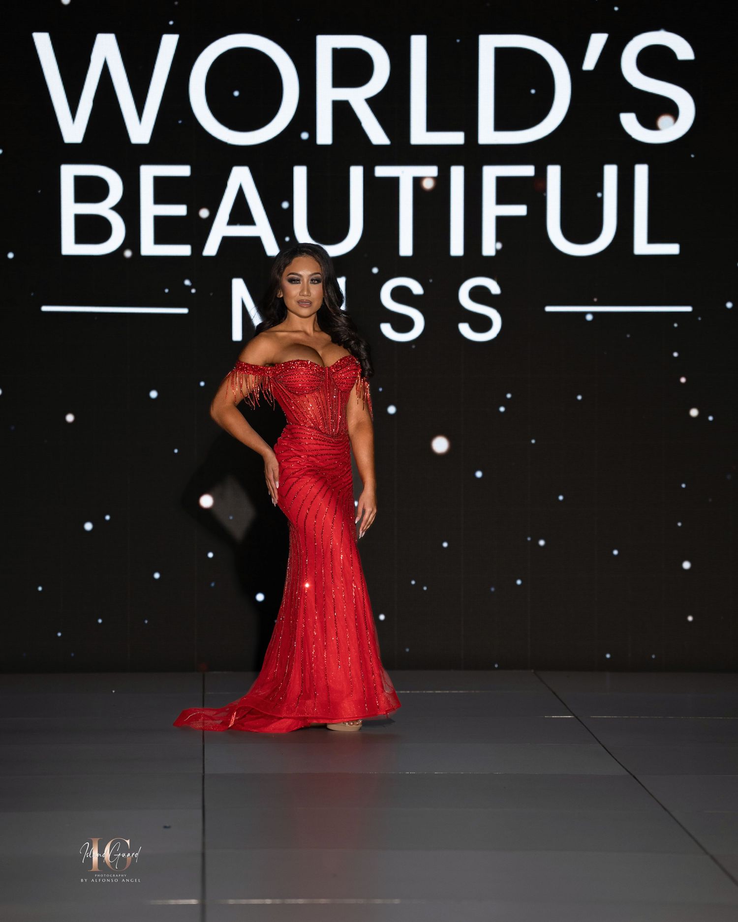 A pageant contestant in a flowing red evening gown poses gracefully on a dark stage with sparkly backdrop.