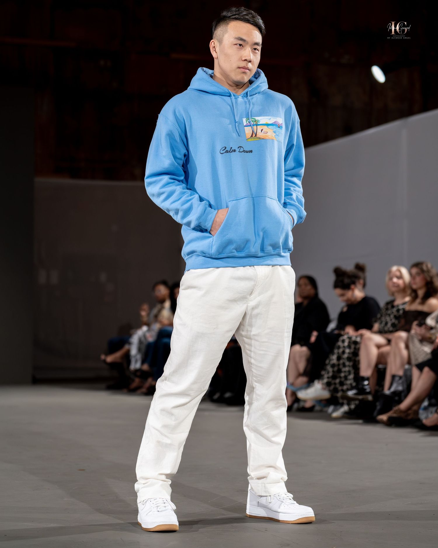 Model walks runway wearing light blue hoodie with white pants and white sneakers during fashion show.