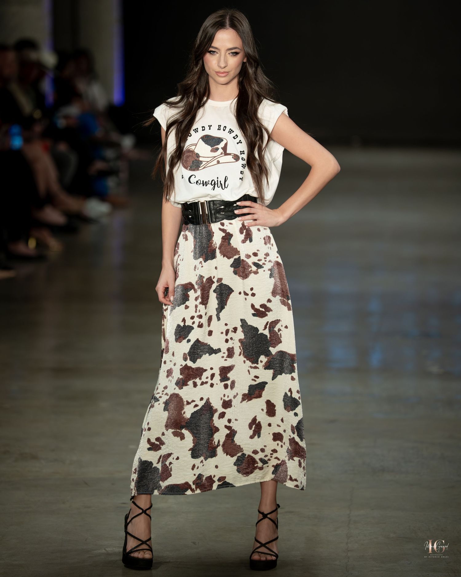 Model walks runway in white t-shirt with brown cow print pattern dress at fashion show.