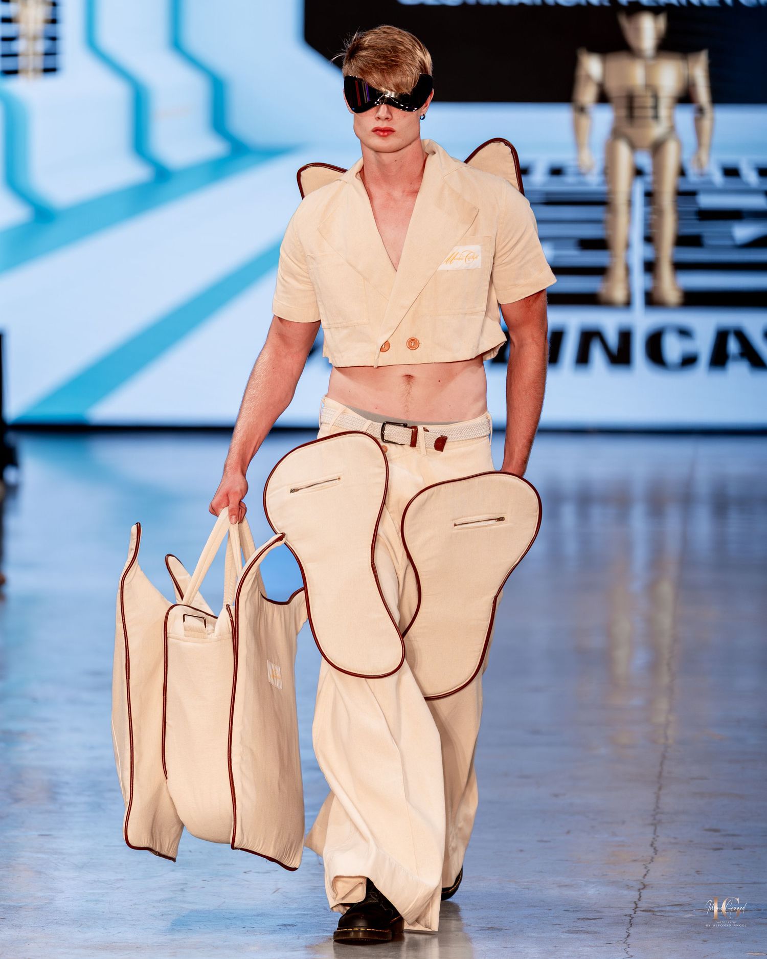 Model walks runway in cream-colored jumpsuit with matching oversized tote bag at fashion show.