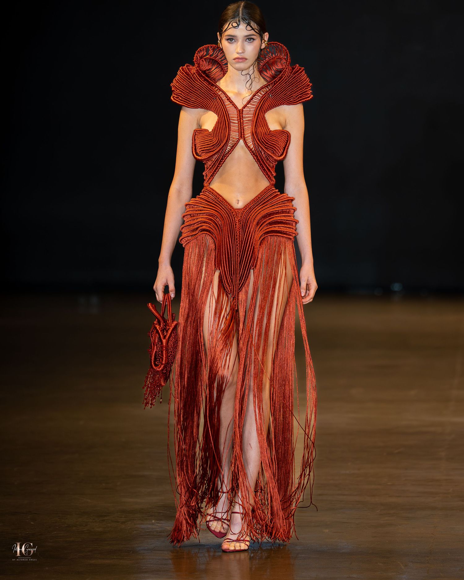 Model wearing a dramatic burnt orange leather dress with sculptural ruffles and fringe details on a dark runway.