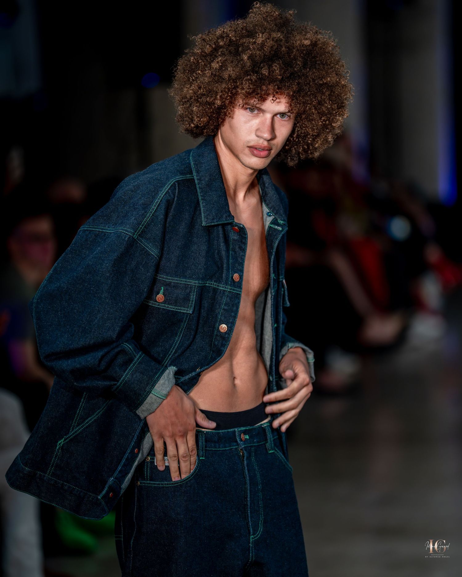 Model walks runway in dark denim outfit with curly hair during fashion show.