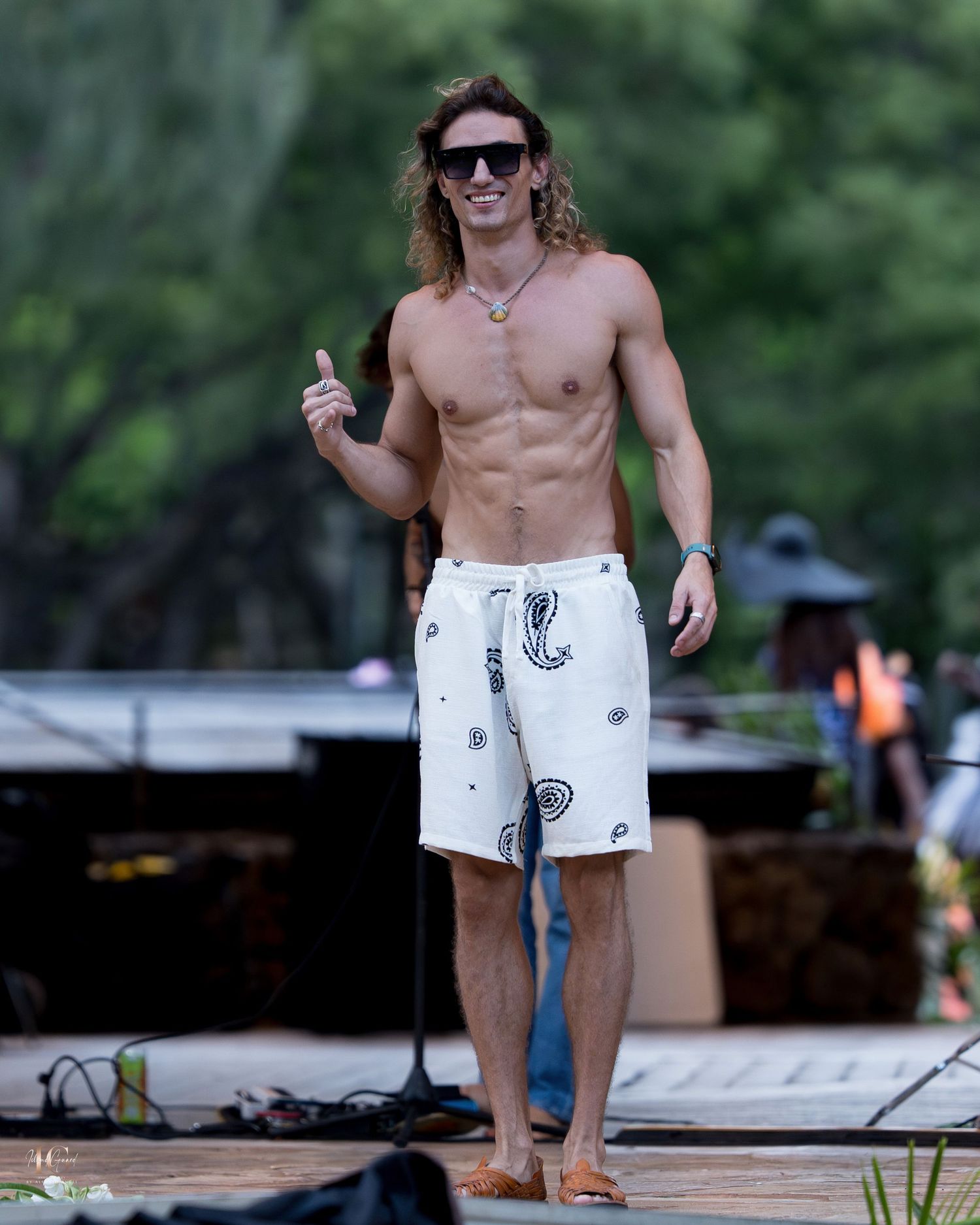 Athletic person in sunglasses and swim trunks stands barefoot on wooden deck outdoors with lush greenery in background.