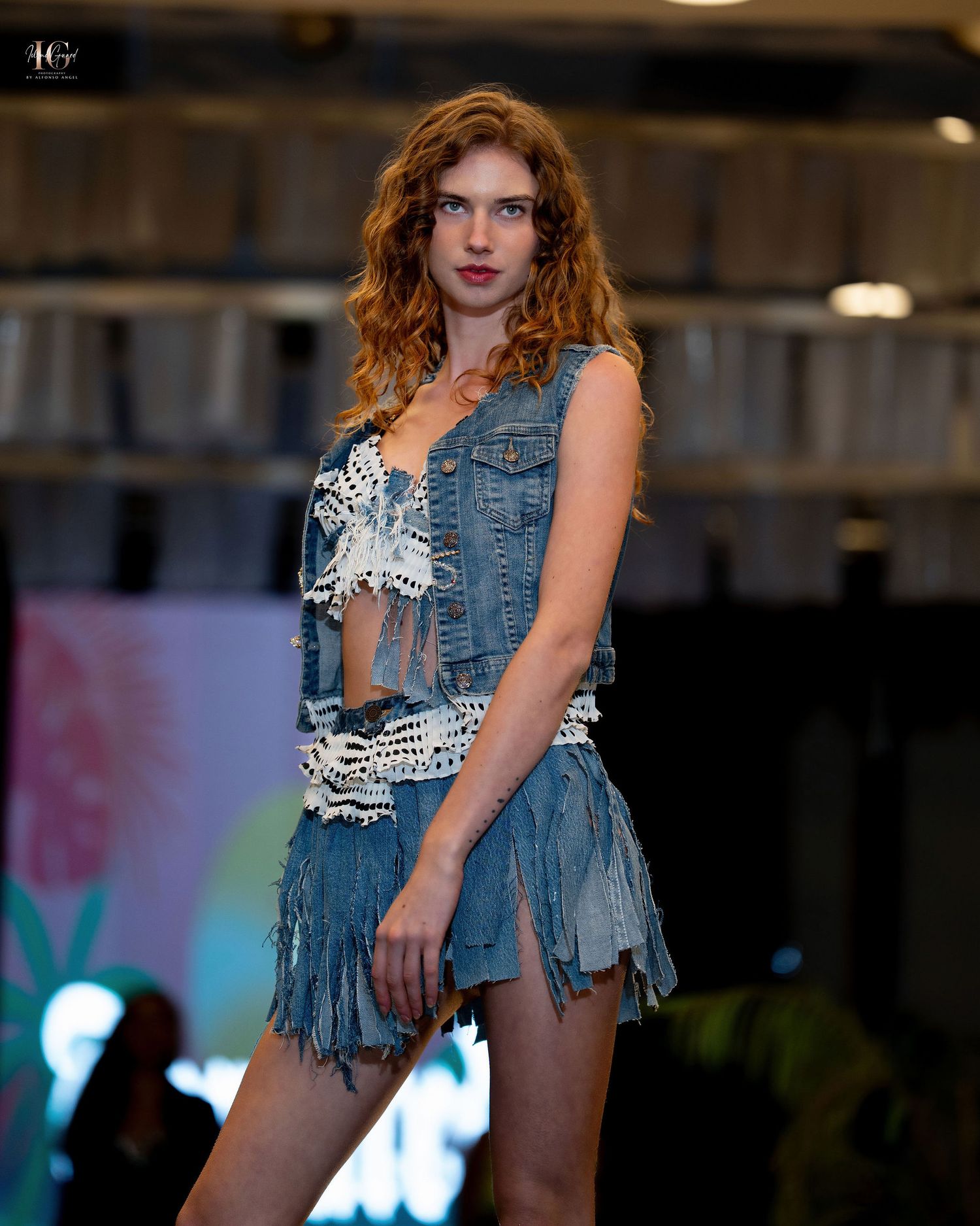 Model walks runway in frayed denim vest and shorts during fashion show with colorful lighting effects in background.