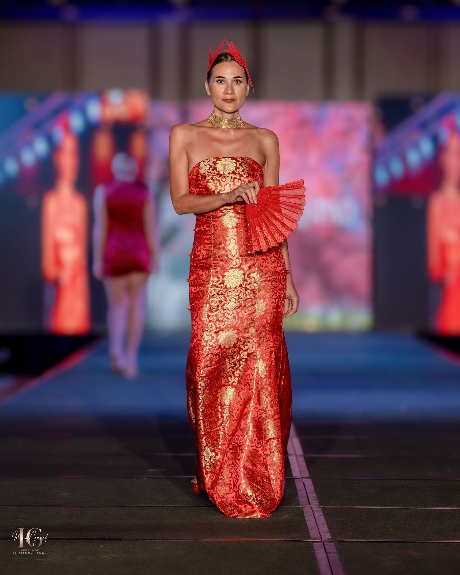 Model presents shimmering red and gold evening gown with matching fan on fashion runway.