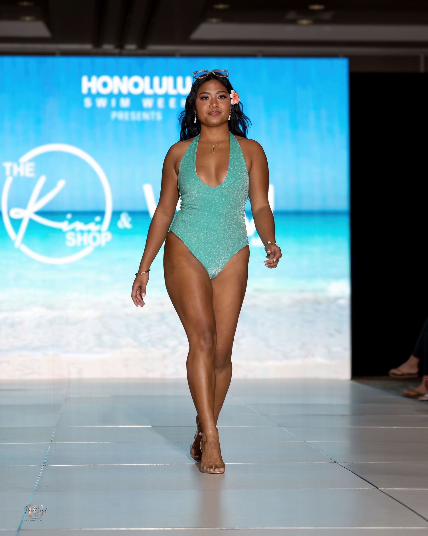 Model walks runway in mint green swimsuit at beachside fashion event.