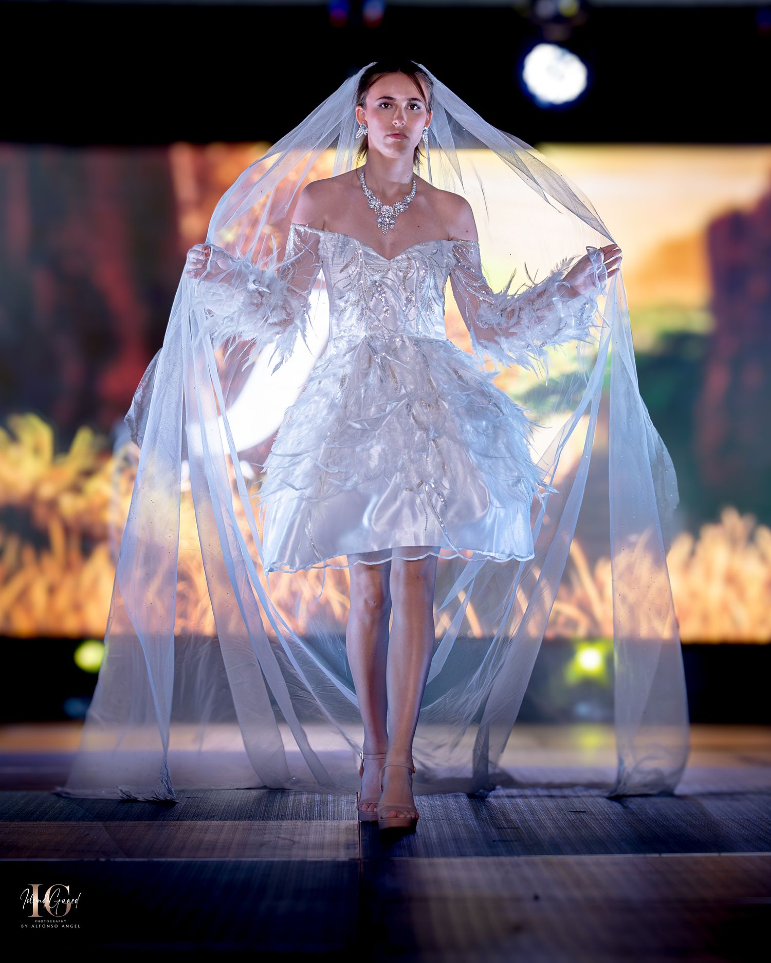 Short white bridal dress with dramatic veil and sheer details on illuminated catwalk.