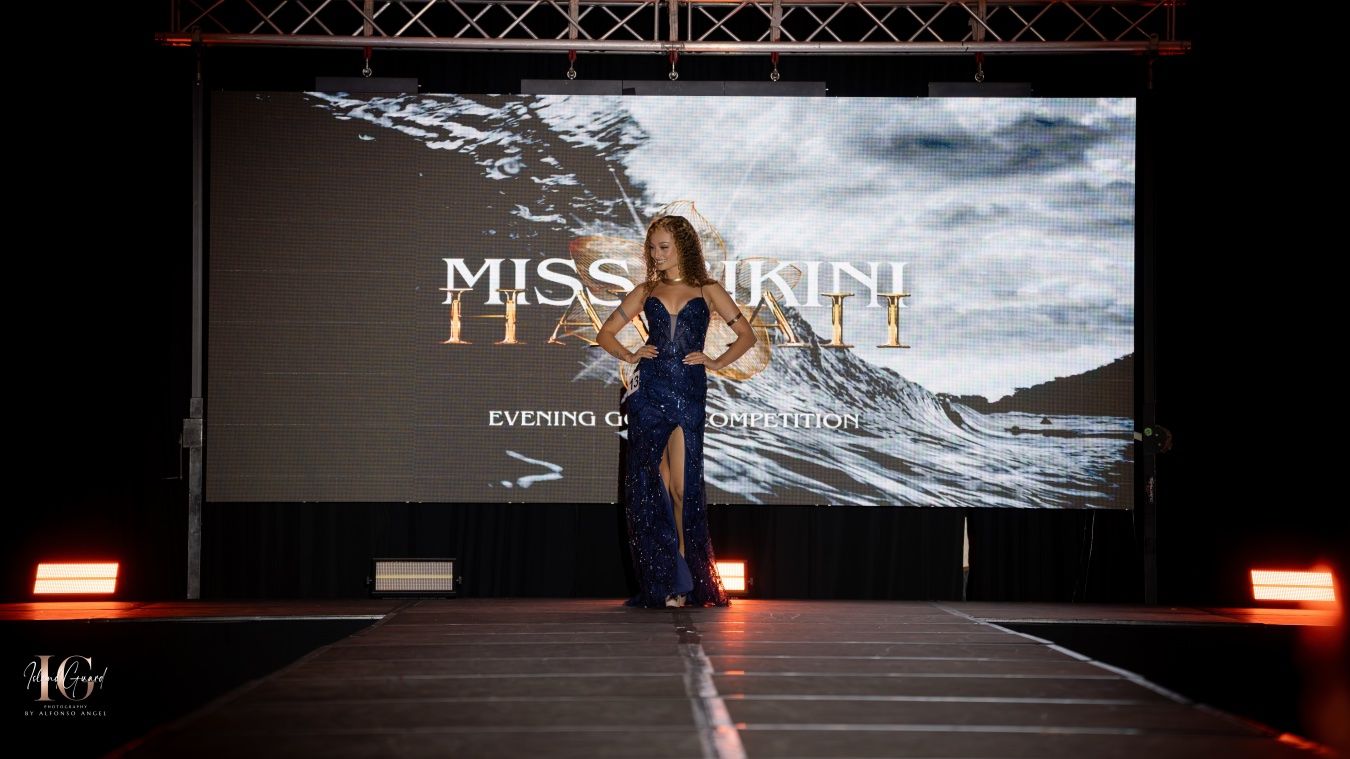 Model walks the runway in evening wear during the Miss Earth fashion show on a dark stage with illuminated backdrop.