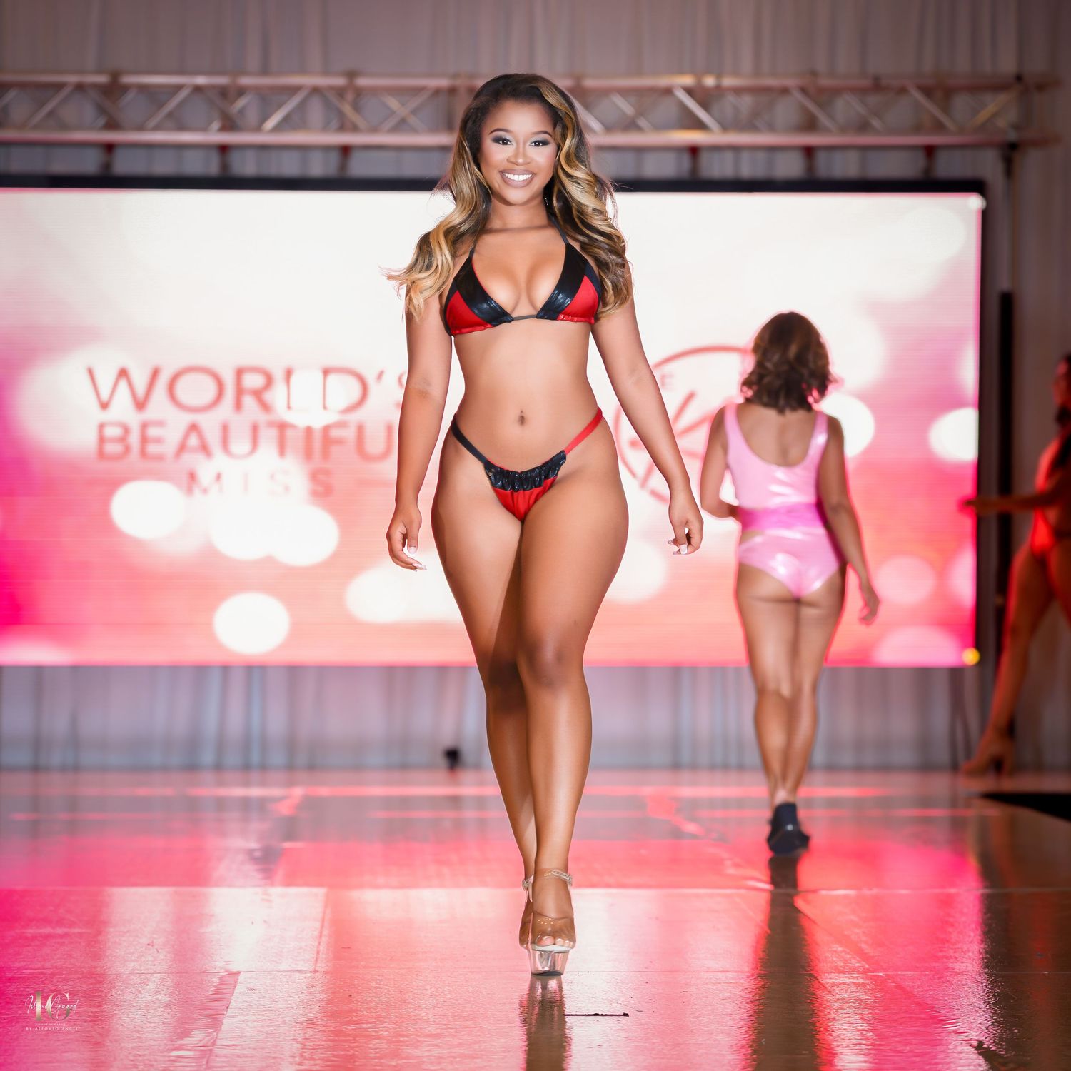 Models walk the runway in red and black swimwear at a fashion show with pink lighting and staging.