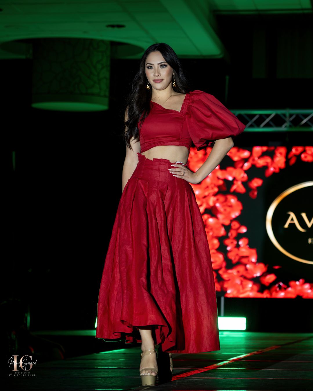 Dramatic red two-piece gown with puff sleeve and flowing skirt showcased on illuminated runway with floral backdrop.