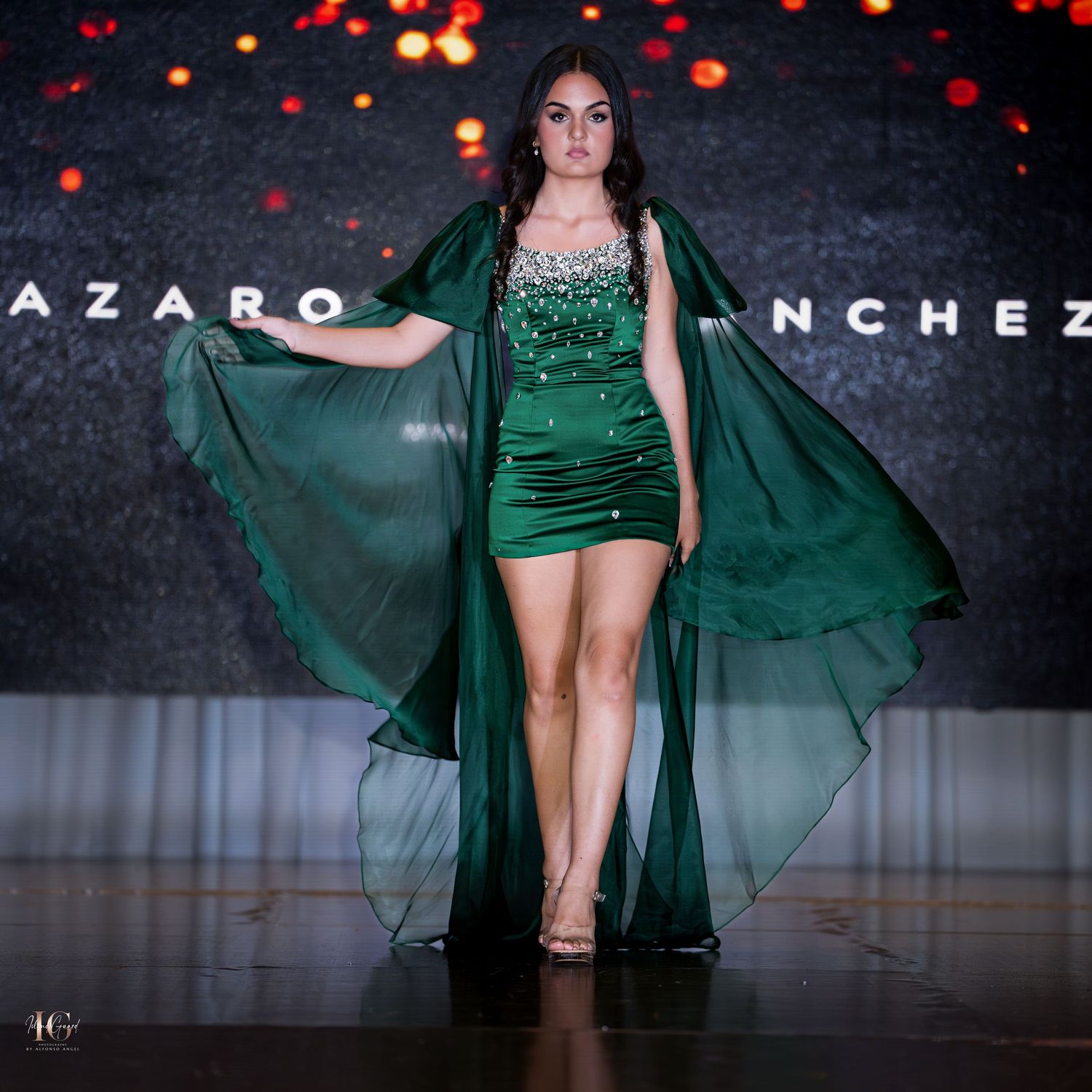 Model presents emerald green mini dress with matching flowing cape on dark runway.