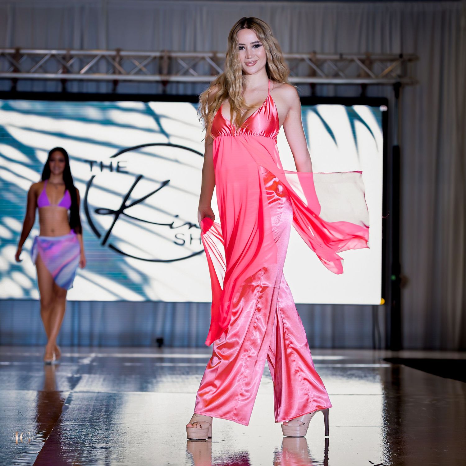 Model walks runway in flowing coral pink jumpsuit at fashion show.
