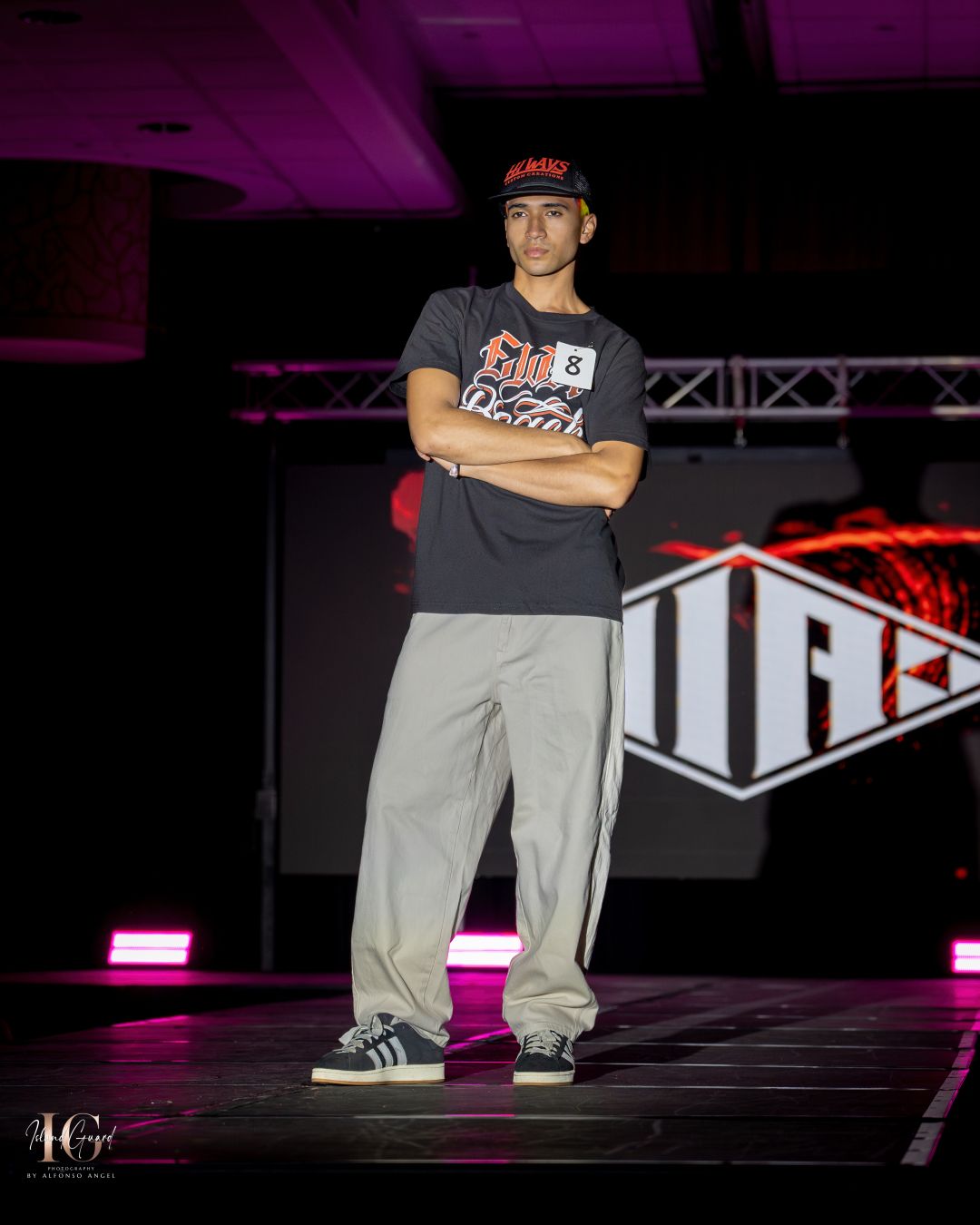 Model in black graphic tee and loose pants poses on illuminated catwalk.