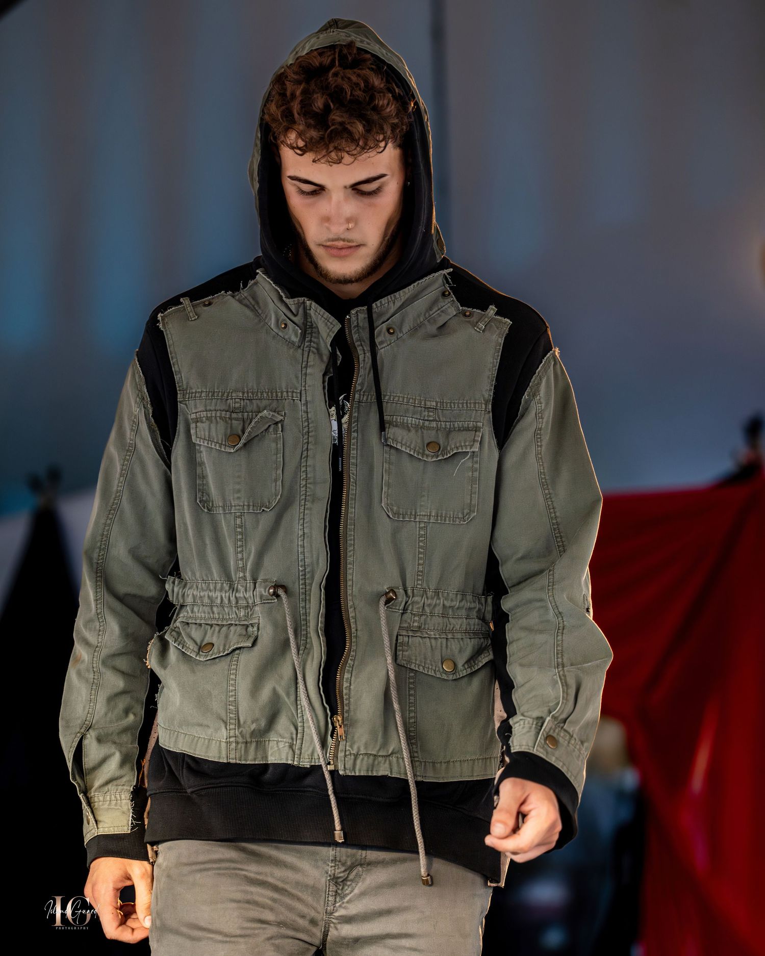 Close-up of model in hooded military-style jacket on dark stage.