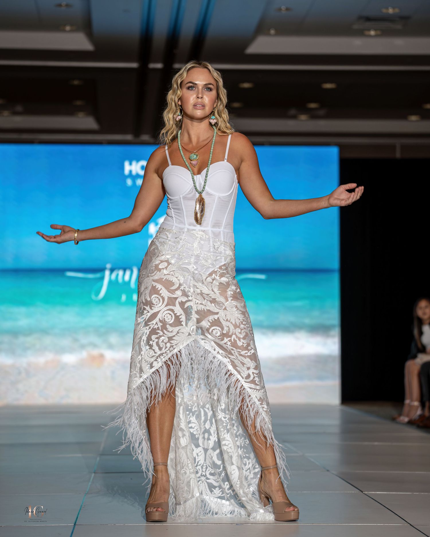 Model presents white lace dress with fringe details at tropical-themed fashion event.