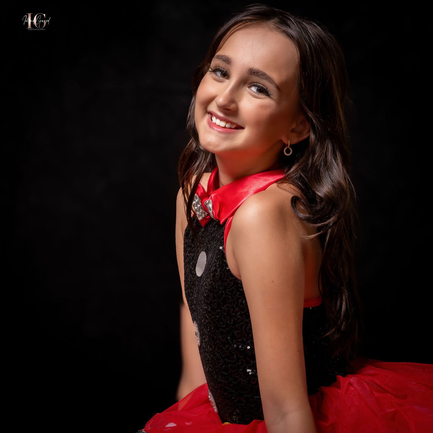 Young dancer in black and red formal dress poses gracefully on dark background.