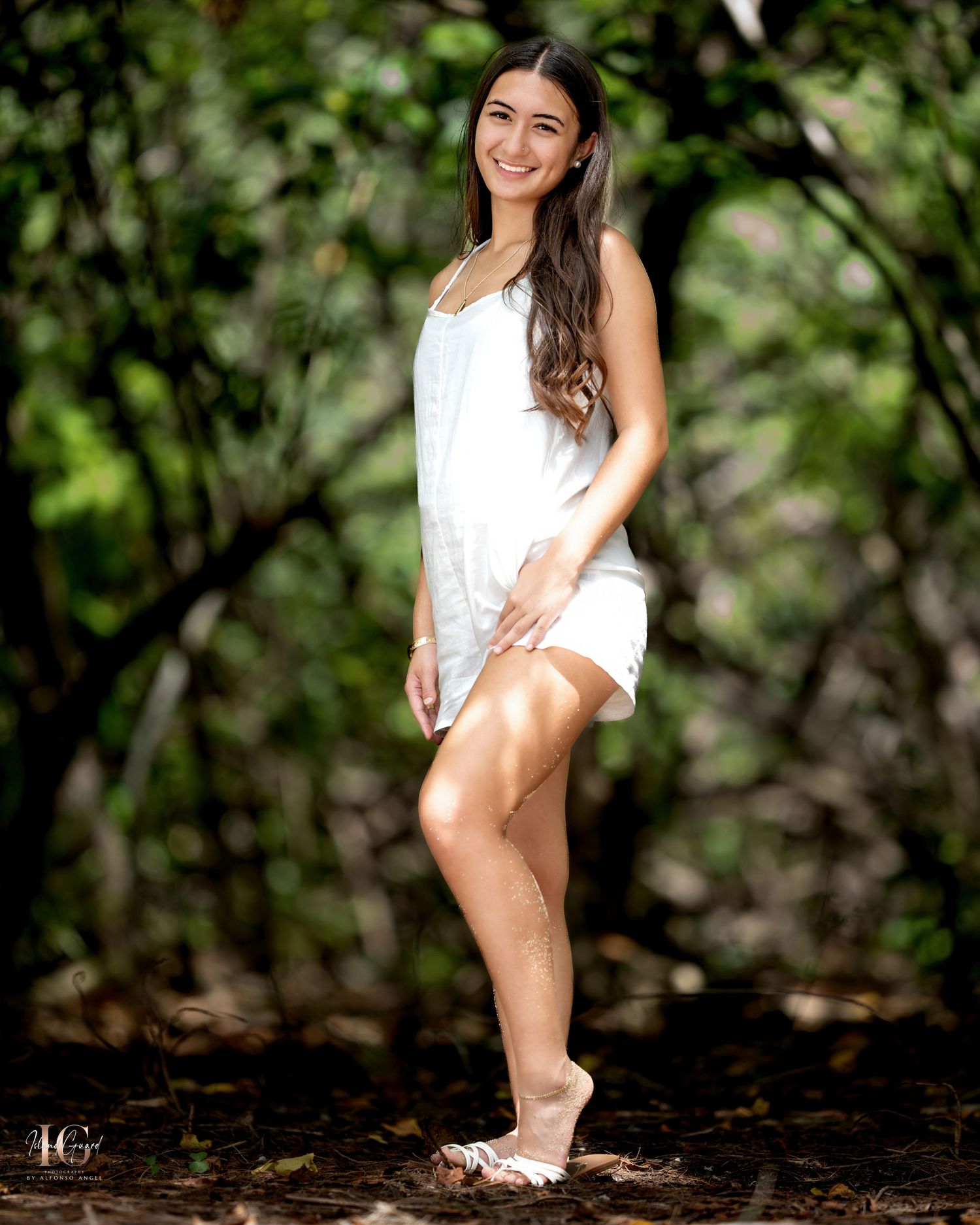 Young woman in white sundress and sneakers poses outdoors in natural forest setting on dark soil.