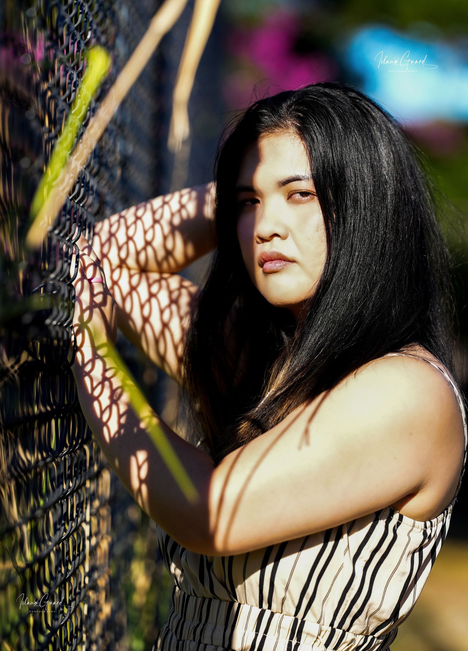 Artistic portrait with dramatic lighting against a chain link fence showcasing long dark hair and striped top.