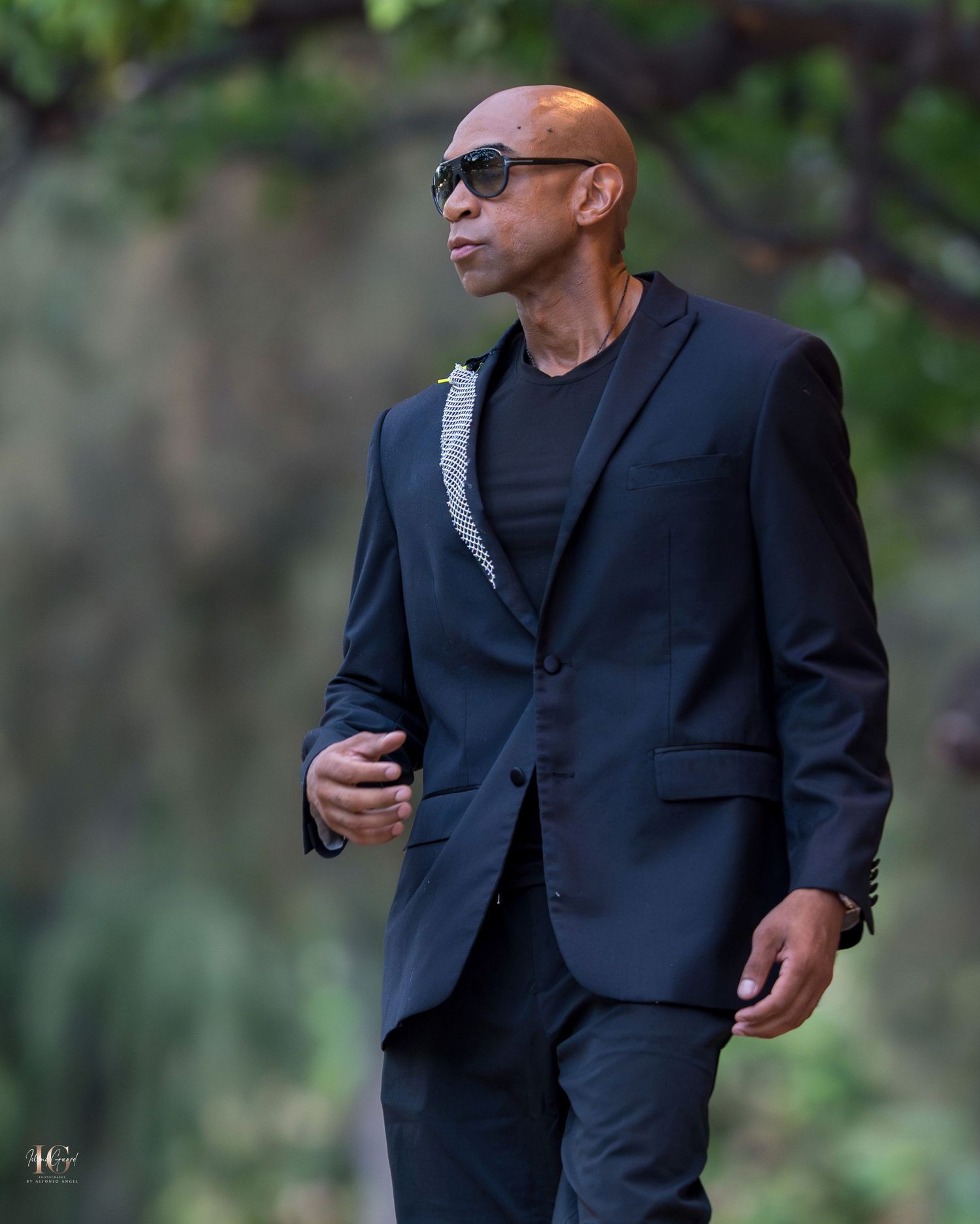 A person in a dark suit and sunglasses walks through a wooded area in a casual manner.