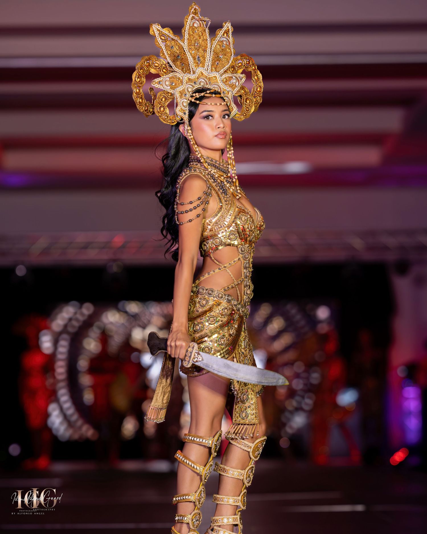 Model in gold warrior costume with sunburst crown poses with sword prop on runway.
