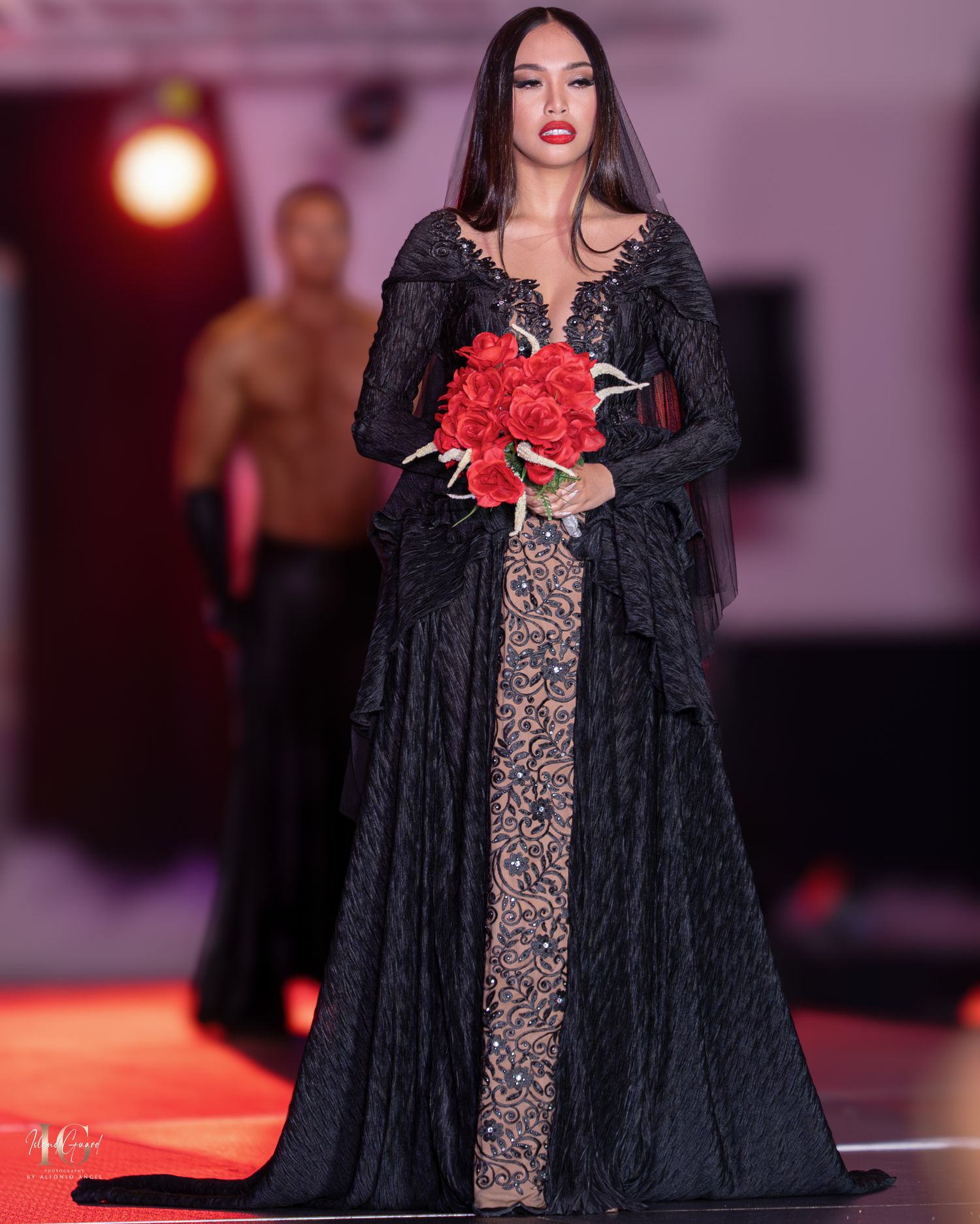 Model walks runway in dramatic black lace gown with red bouquet at fashion show.