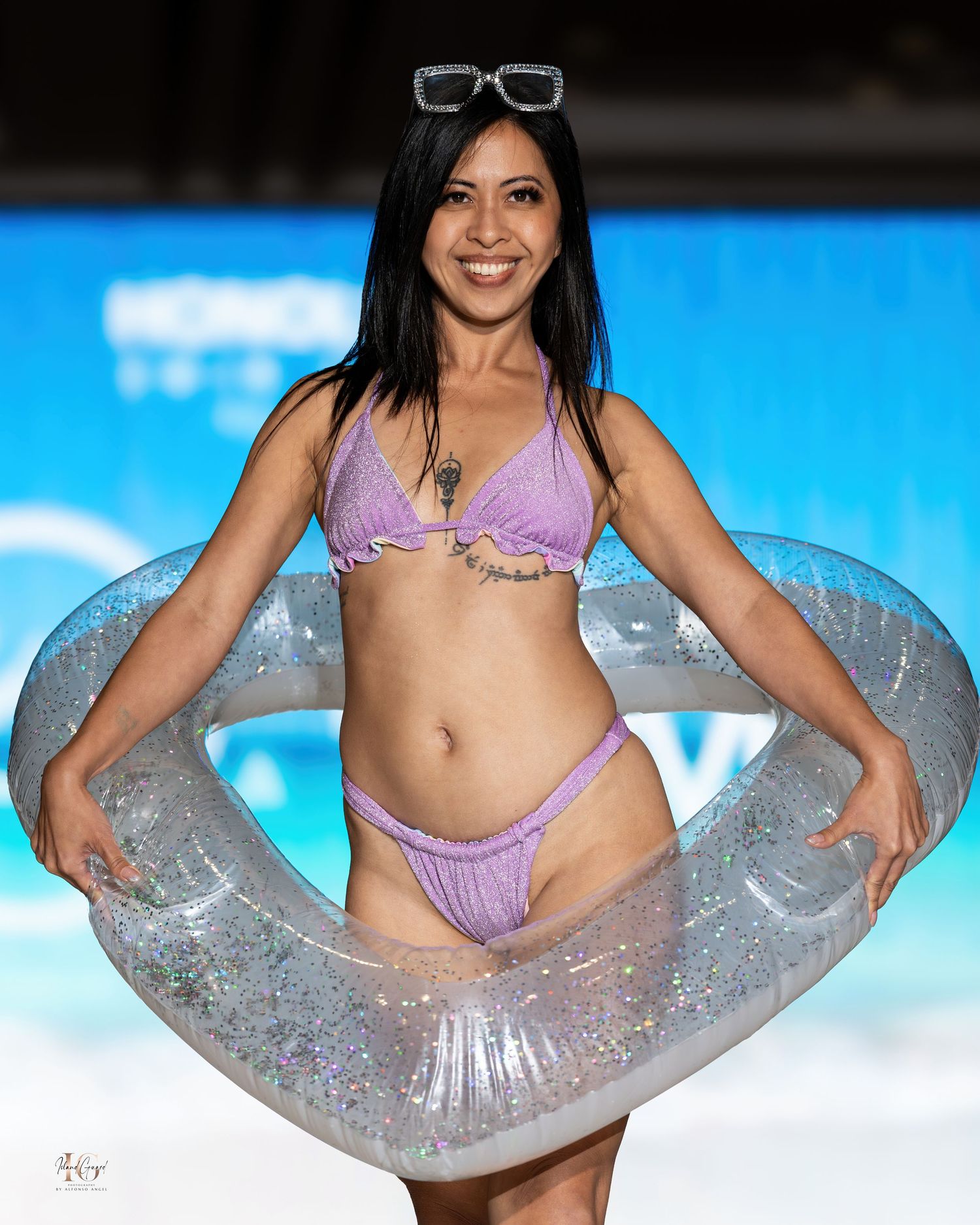 Model showcases purple bikini while holding transparent pool float on catwalk.
