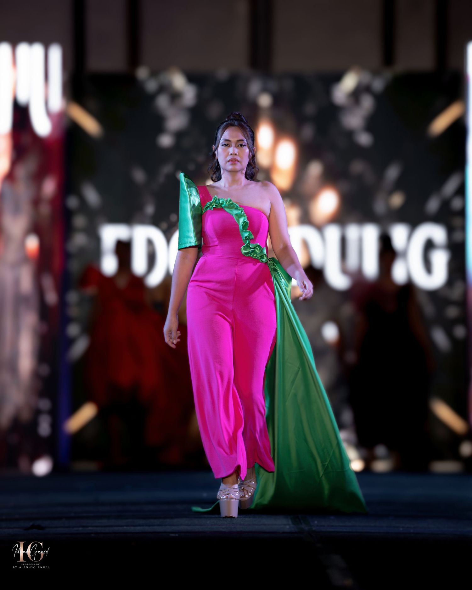 Fashion model wearing flowing pink dress with green cape moving dramatically on stage.