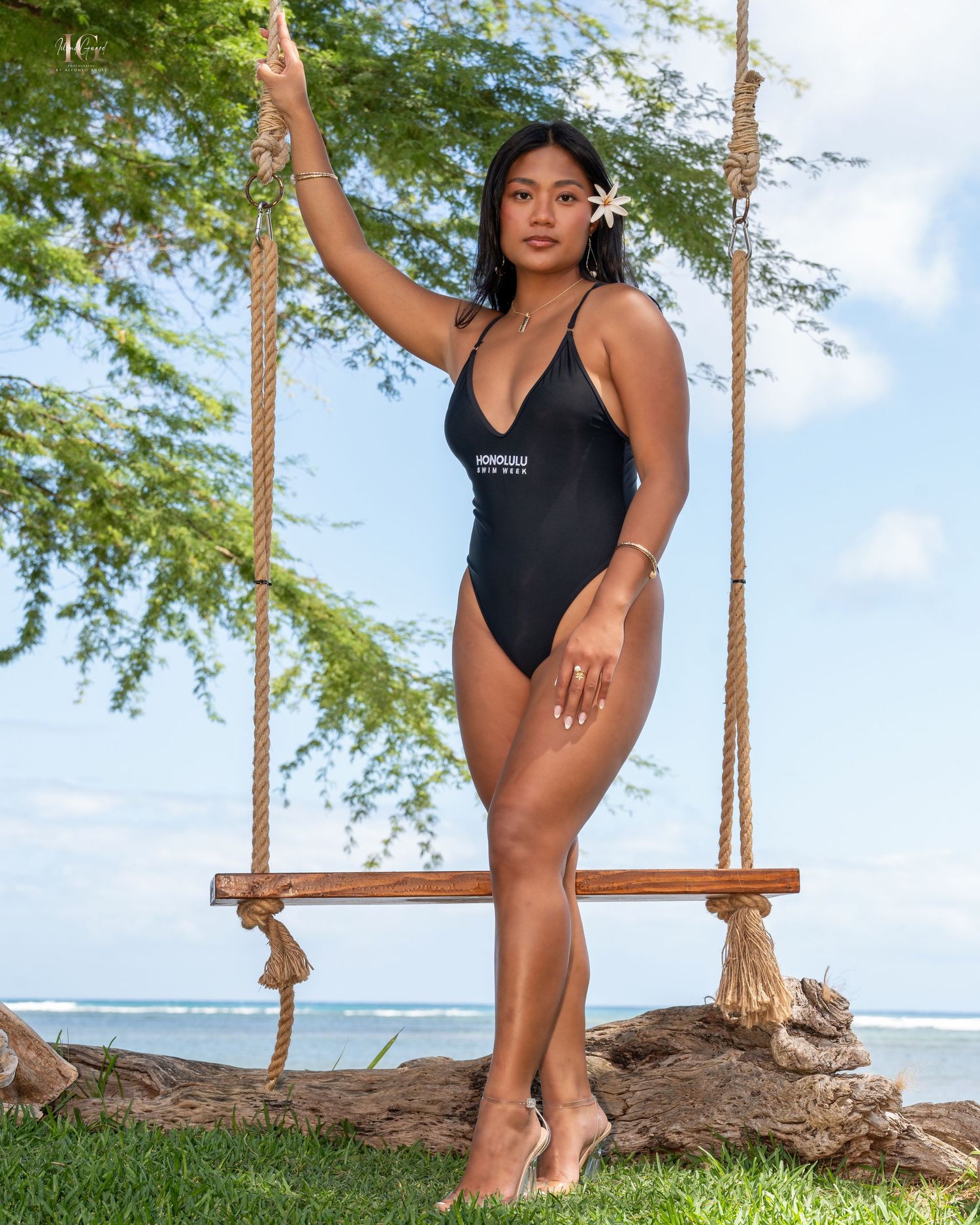 A person in a black one-piece swimsuit poses on a wooden rope swing by the ocean with palm trees in the background.