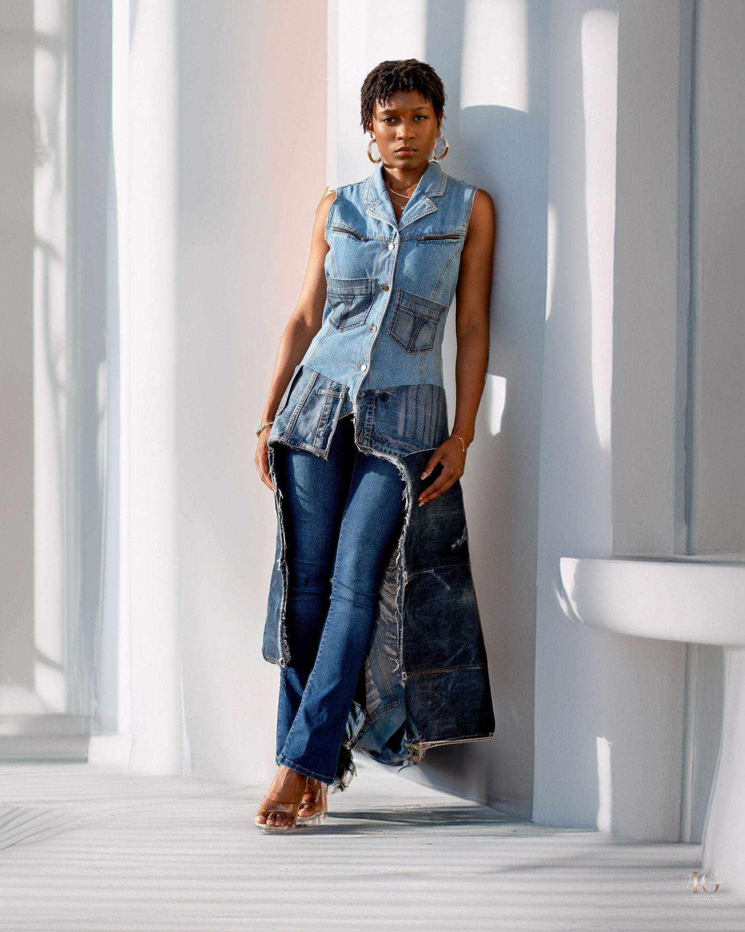 Model posing in trendy denim outfit with sleeveless jacket and jeans against white wall in natural light.
