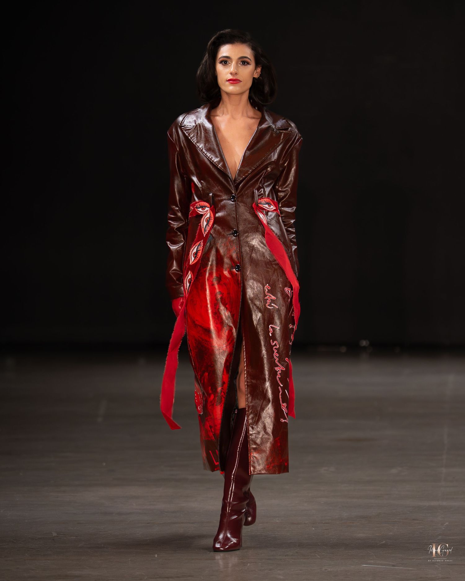 Model walks runway in burgundy patent leather trench coat with red accents during fashion show against dark background.