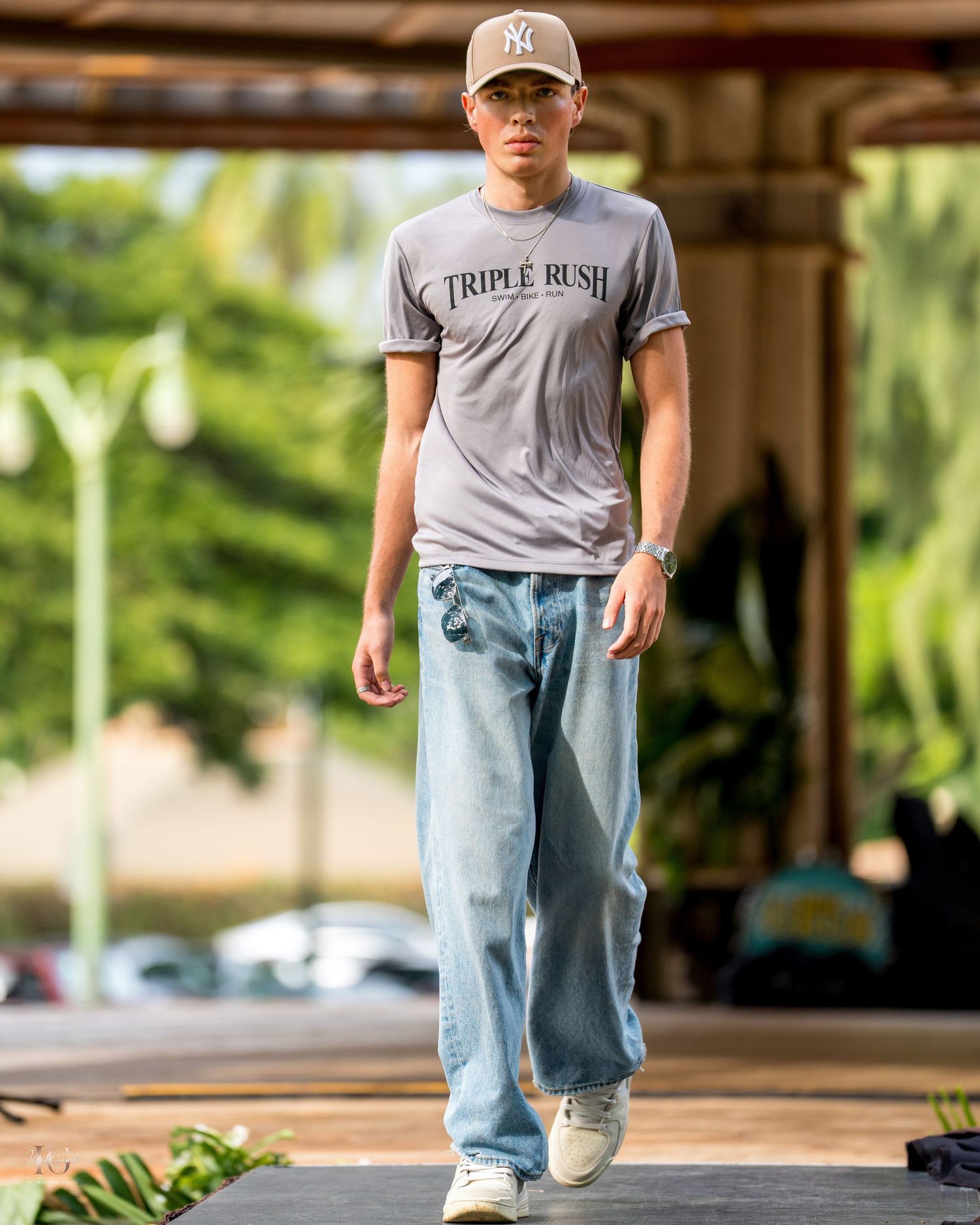A person wearing a gray t-shirt and light blue jeans walks on a wooden deck with palm trees in the background.