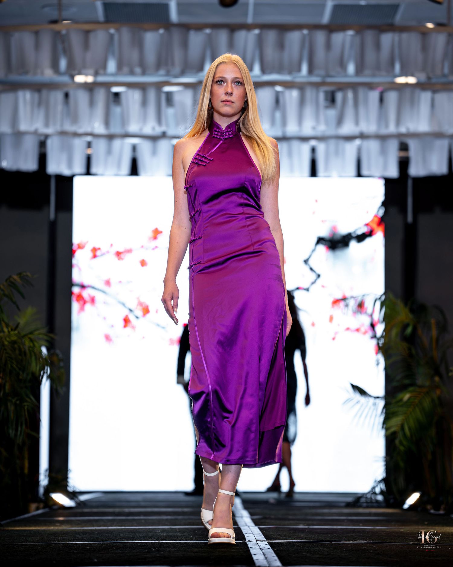 Model in flowing purple evening gown poses on runway with dramatic backlit display and decorative elements.