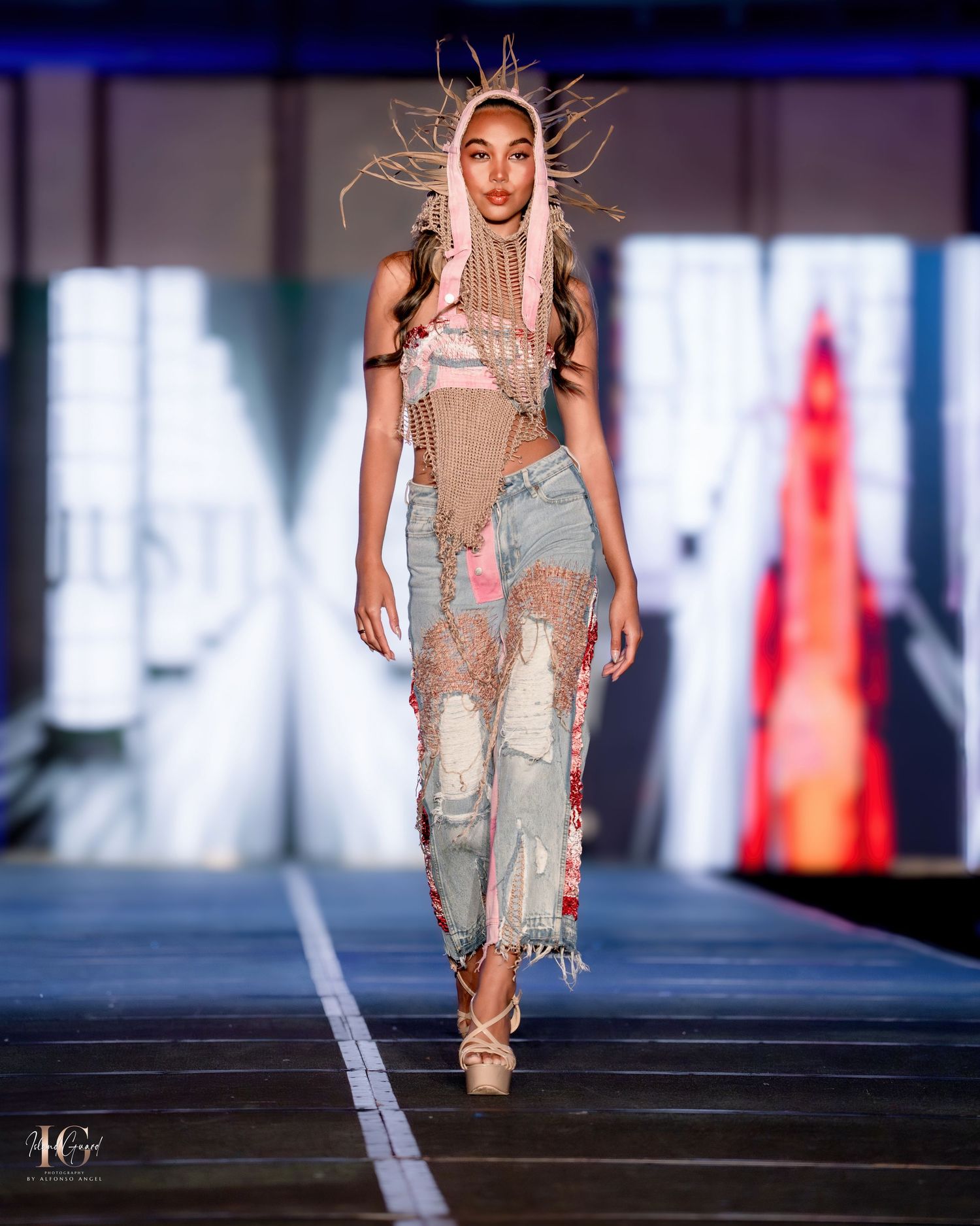 Model wearing beige tattered dress walks runway with dramatic backlighting.