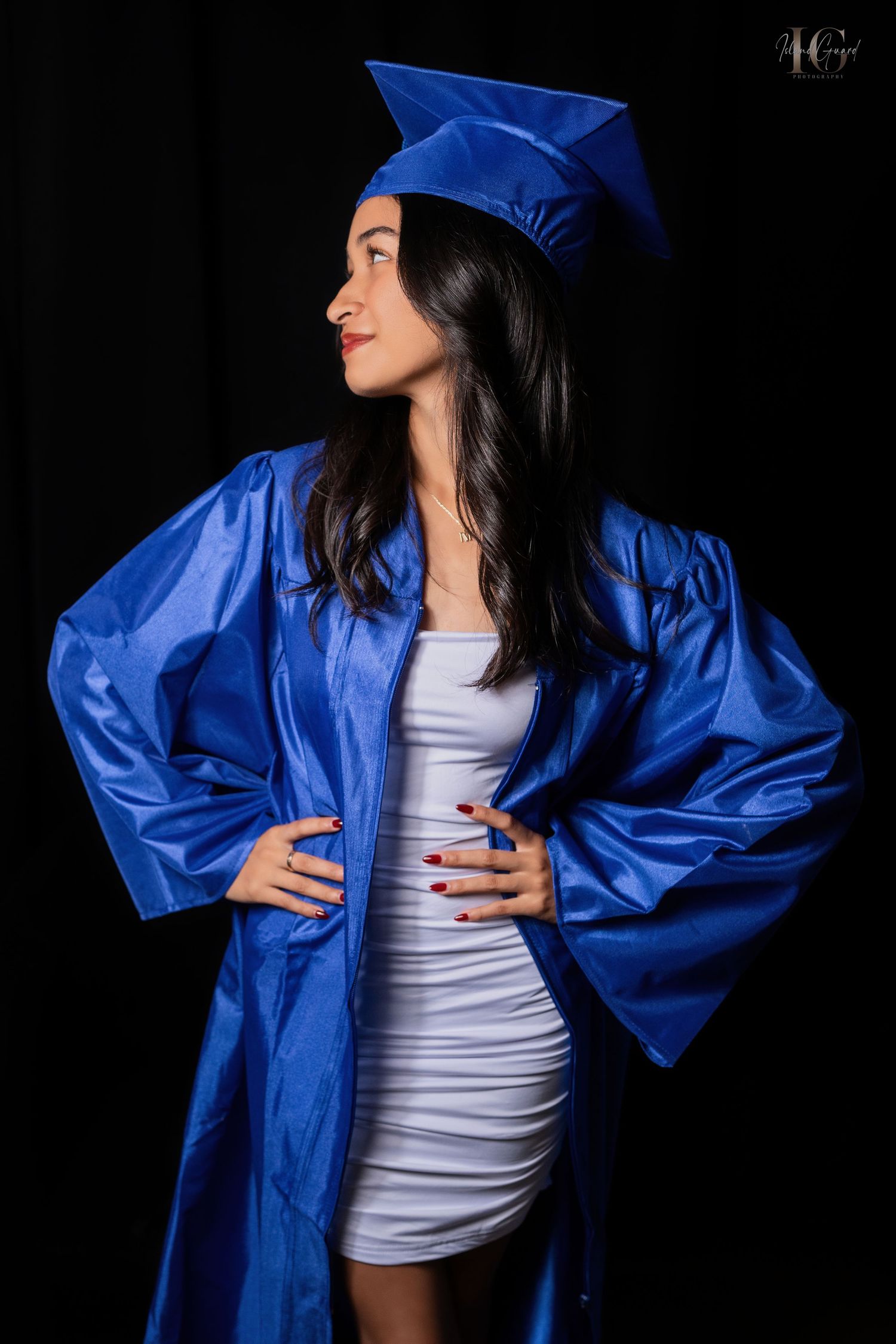 Graduate wearing bright royal blue cap and gown over white dress poses confidently against dark background.