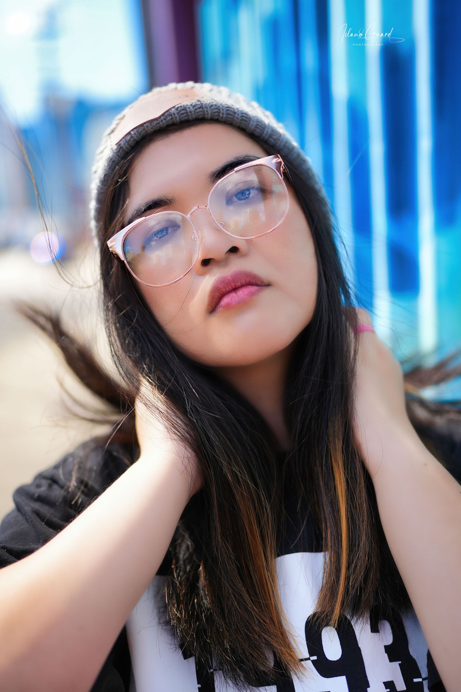 Portrait of someone with long dark hair wearing glasses against a blue-lit background with artistic lighting.