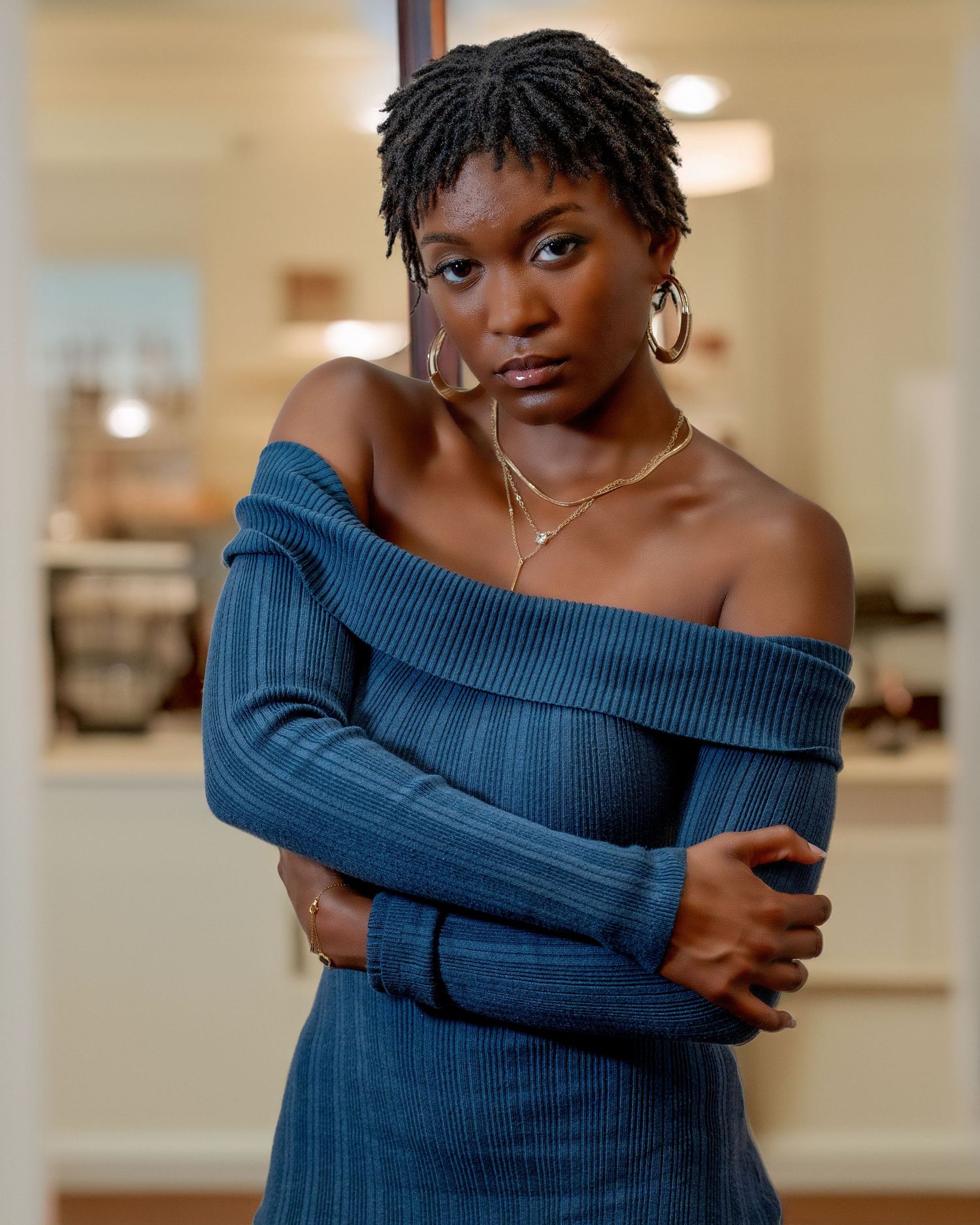 Person in navy blue off-shoulder sweater poses with crossed arms against neutral interior background.