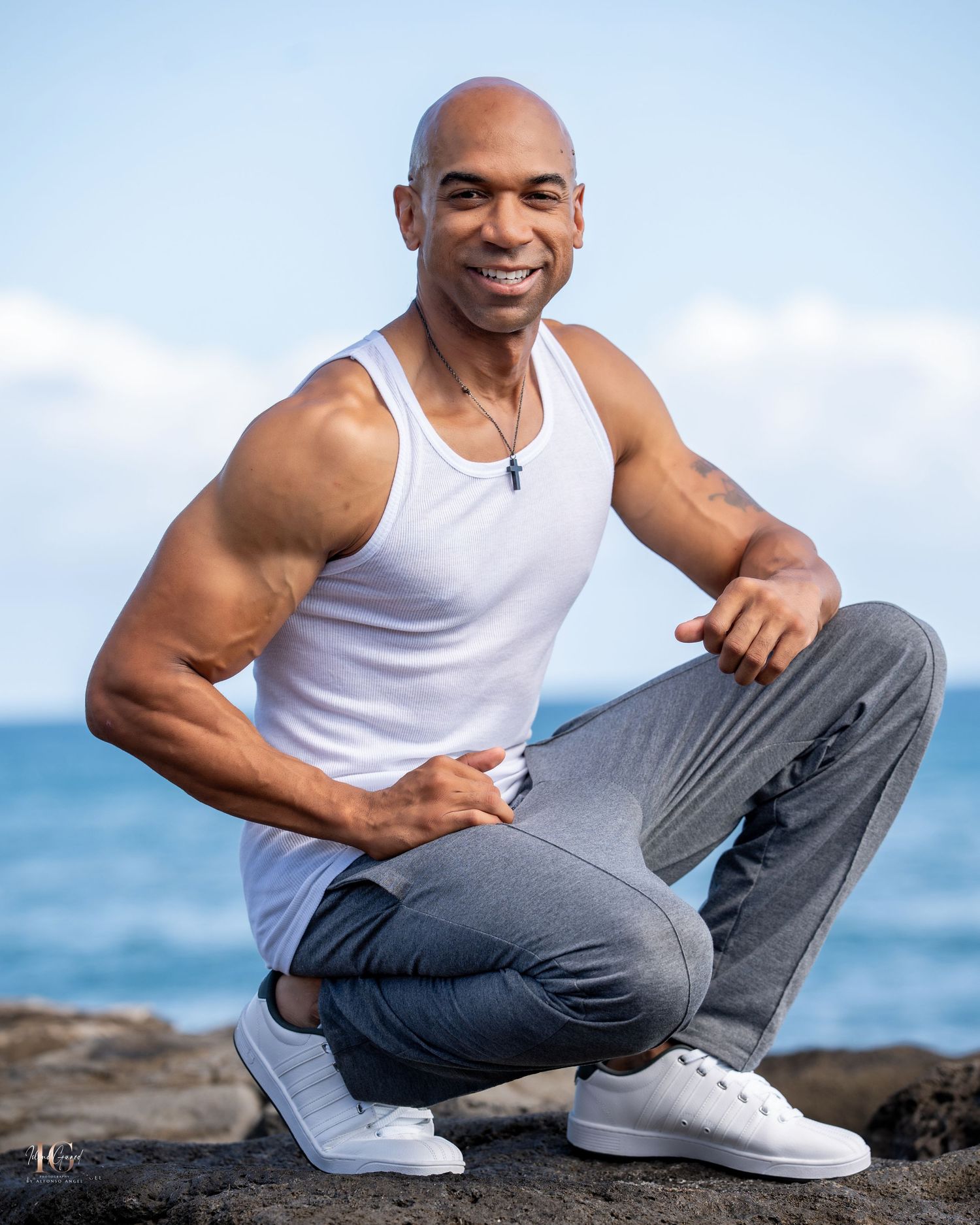 Smiling athletic person in white tank top and gray pants crouches by the ocean on rocky shore during daytime.