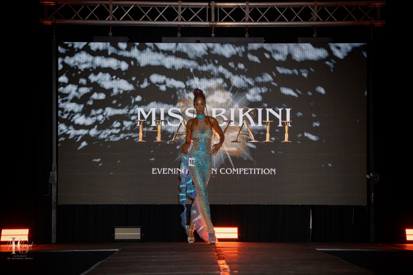 A figure poses on stage in front of a large projection screen displaying 'Miss Bikini' during a competition event.