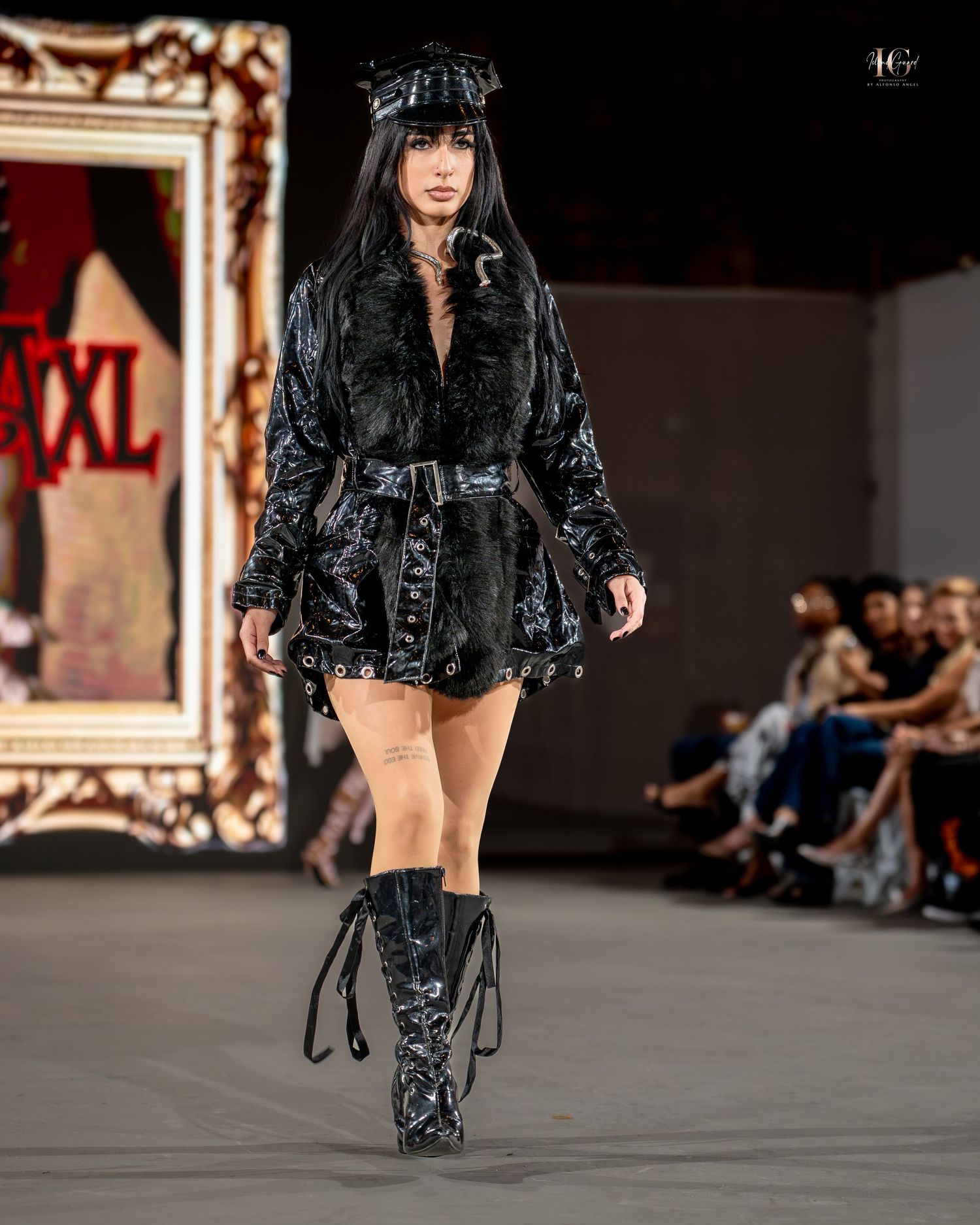 Model walks runway in black shiny coat and patterned pants during fashion show presentation.