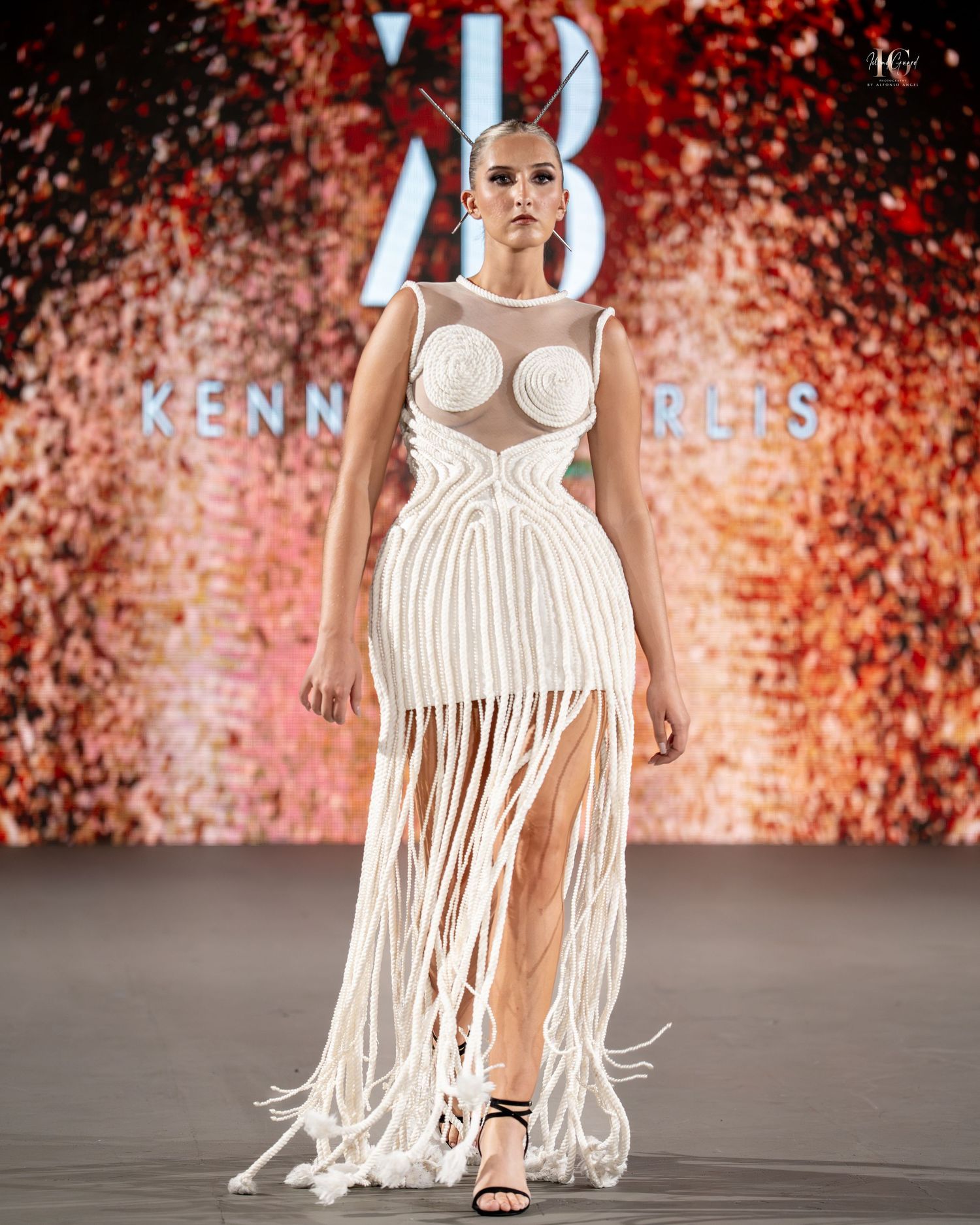 A model walks the runway in a white macrame dress with fringe details and geometric bodice at a fashion show.