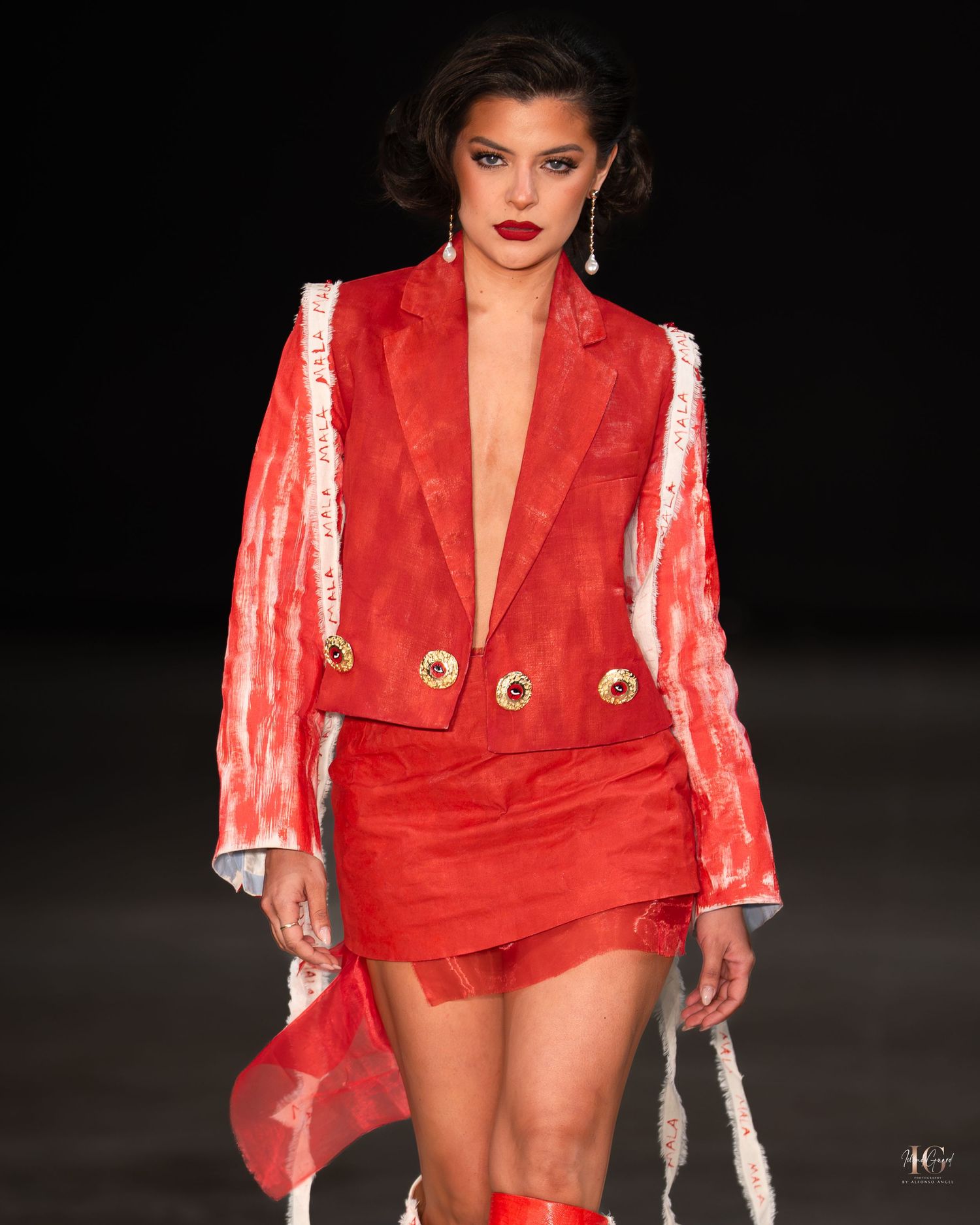 Model walks runway in striking red velvet mini dress with sheer sleeves and gold button details at fashion show.