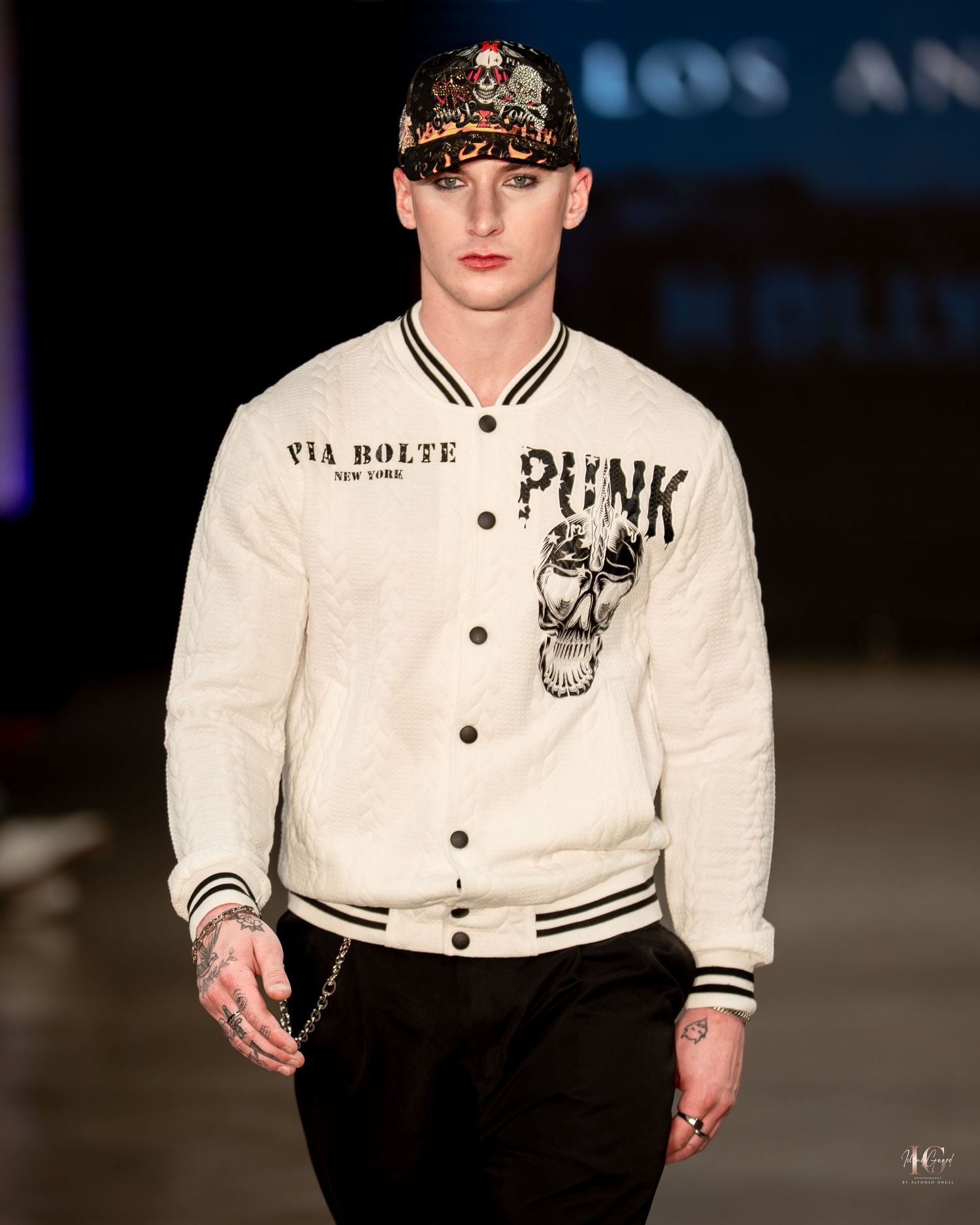 Model on runway wearing white varsity jacket with punk lettering and embellishments during fashion show.