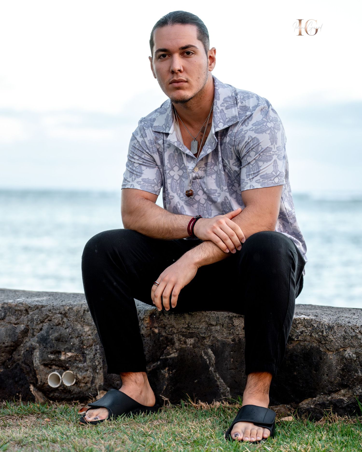 A person in a gray floral shirt sits crouched on a stone wall by the ocean at sunset.