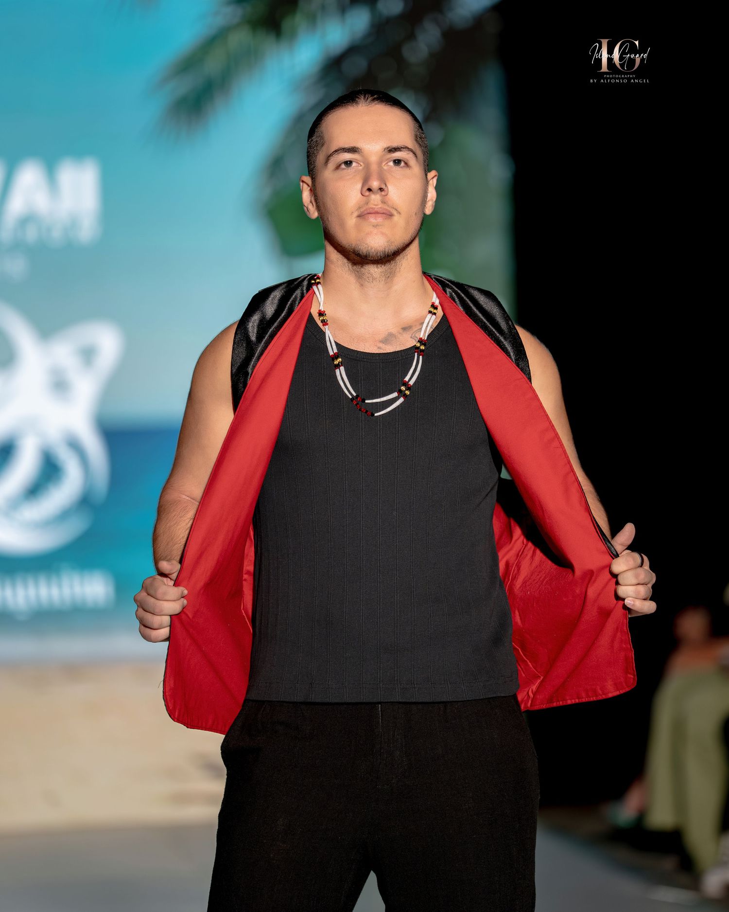Model on runway wearing black tank top and red jacket at beach-themed fashion show with palm trees in background.