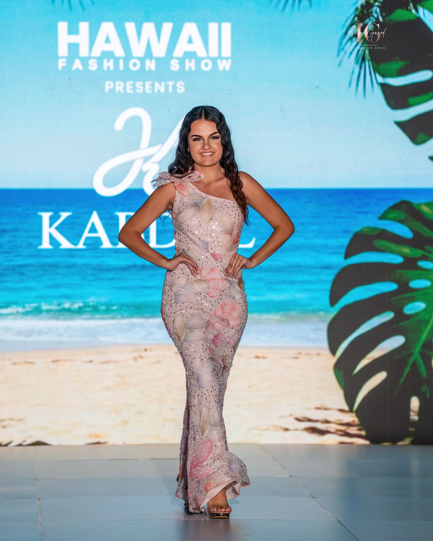 Model walks runway in pink floral gown at tropical beach-themed fashion show with ocean backdrop.